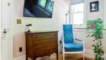 Una habitación con un aparador de madera, un sillón tapizado en azul, un televisor de pantalla plana en la pared, una planta en maceta y una ventana con persianas.