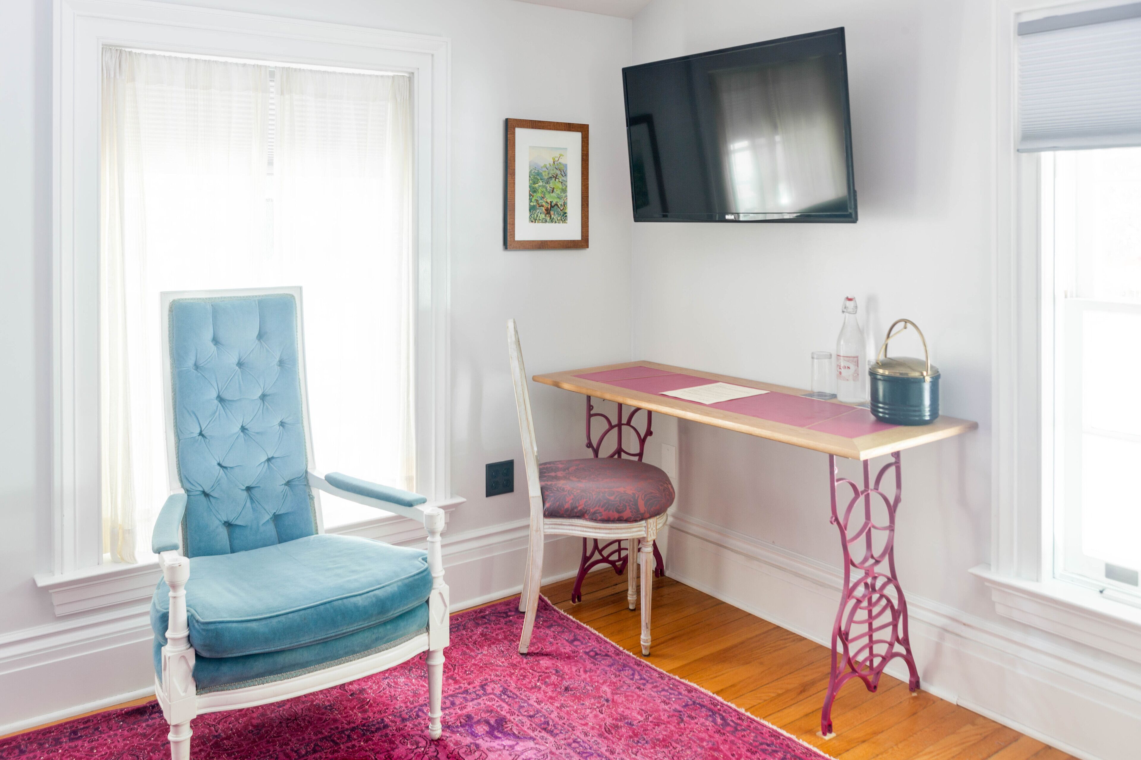 A room with a blue chair, a wooden desk, a pink rug, and a television mounted on the wall.