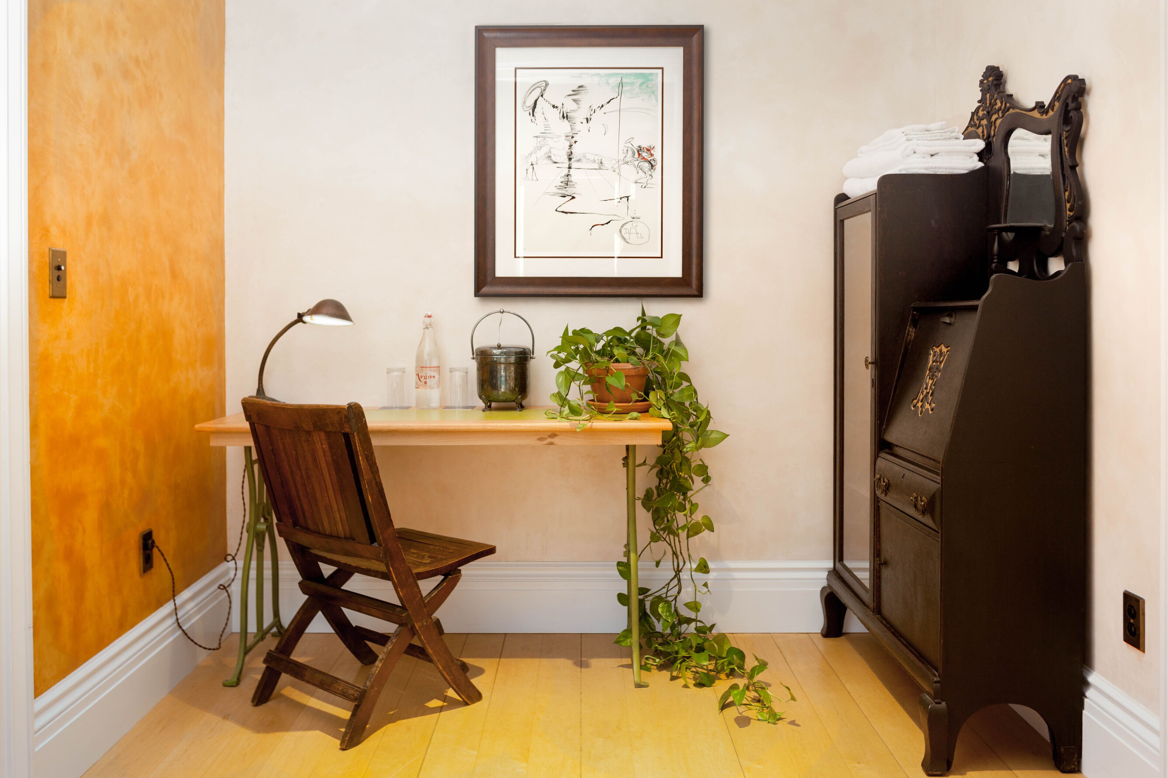 A room with a wooden desk, a chair, a framed artwork on the wall, a cabinet, and a potted plant.