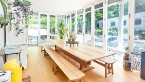 A spacious room with a large wooden table and benches, large windows with a view of the street, and potted plants. - Argos Inn (Ithaca)