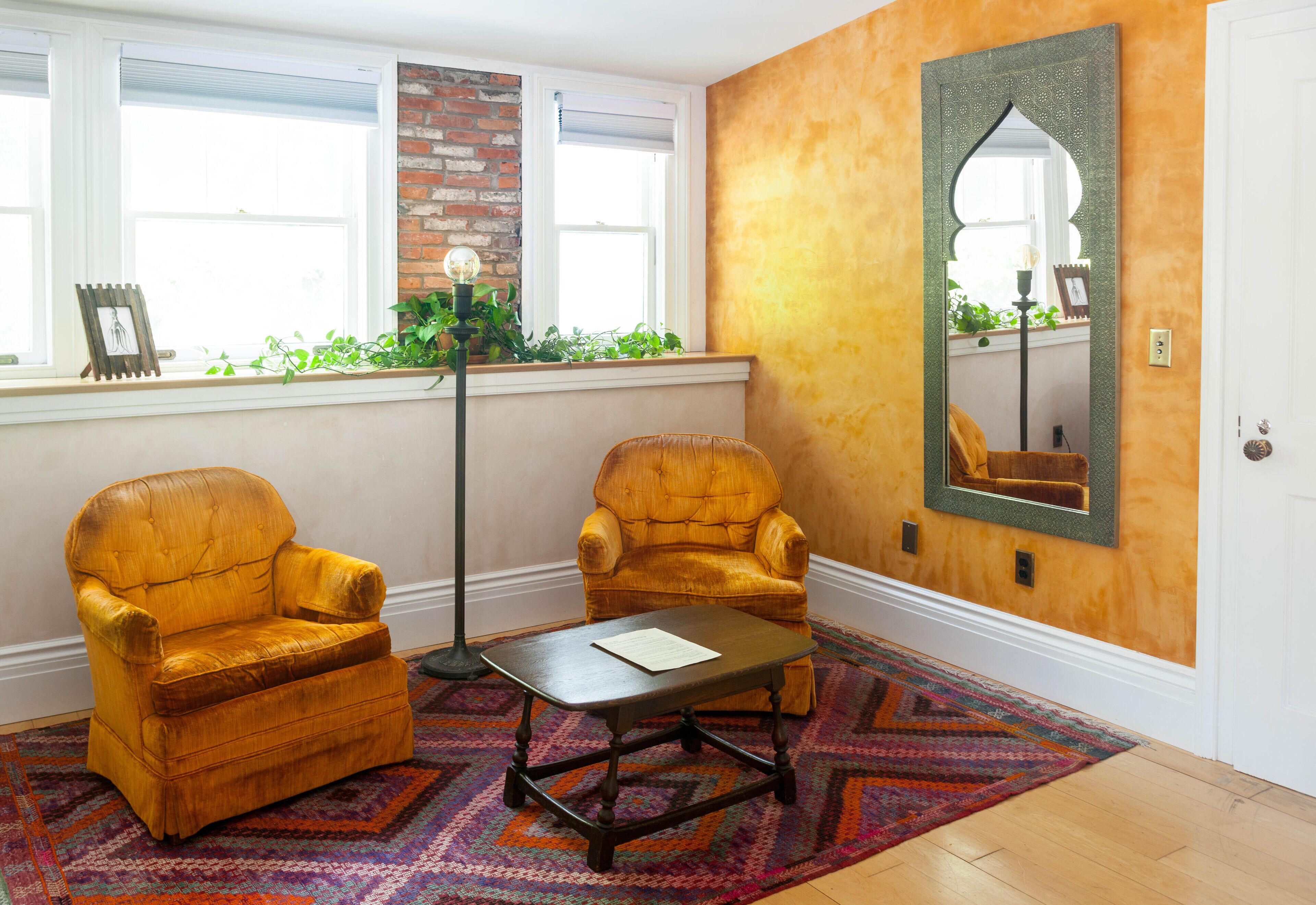 A room with two yellow armchairs, a small table, a patterned rug, a floor lamp, and a large mirror on the wall.