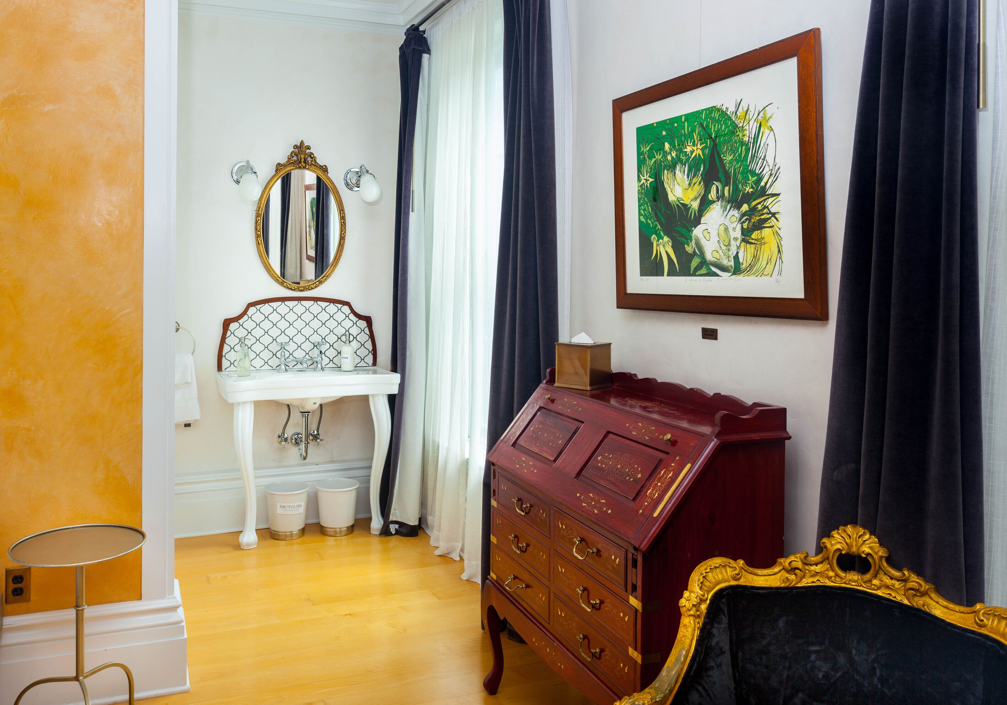 Una habitación con un lavamanos blanco sobre pedestal, un aparador rojo con cajones, un cuadro enmarcado y una butaca tapizada de color negro.