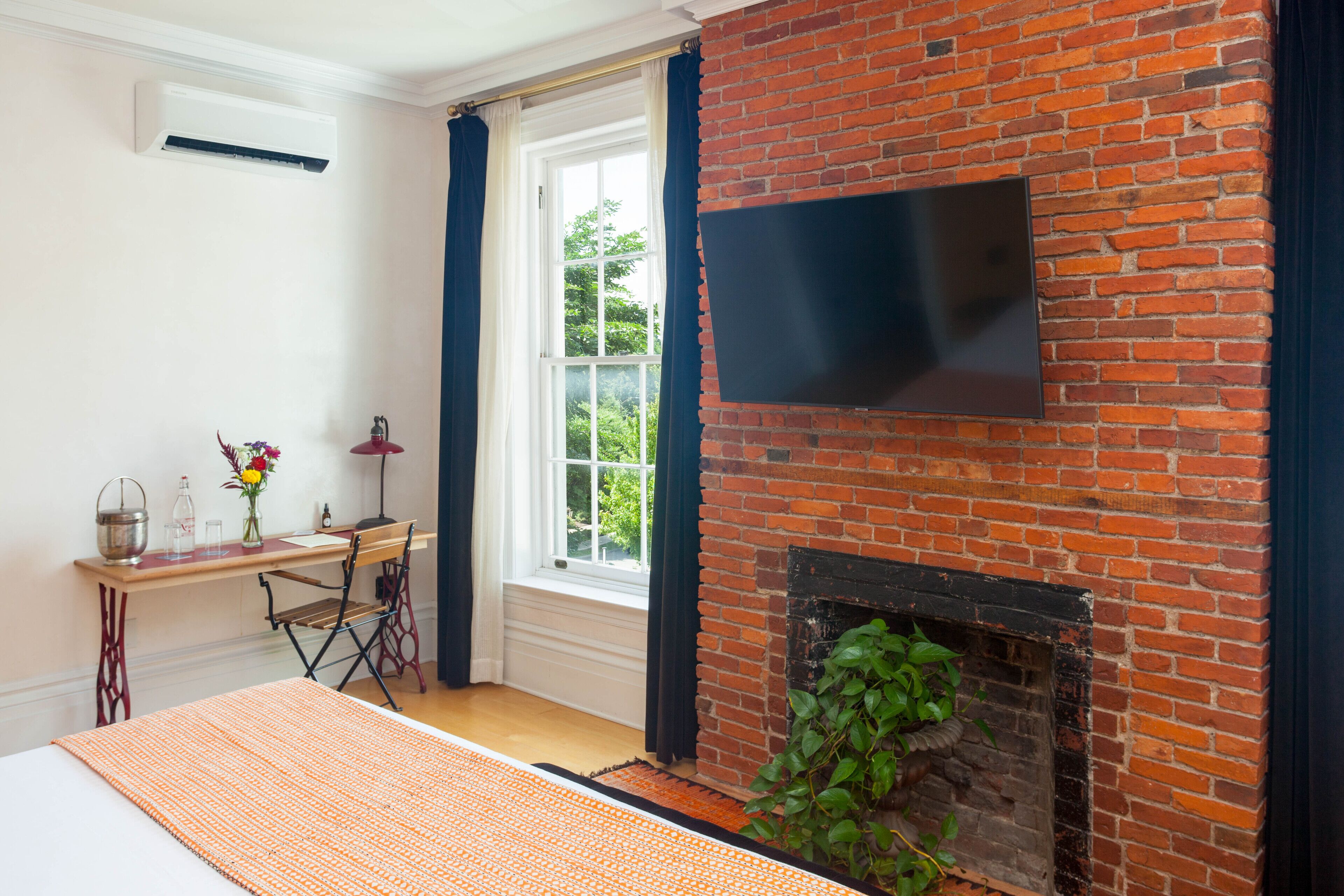 A room with a brick fireplace, a flat-screen TV mounted above it, a desk with a chair, and a window with curtains.