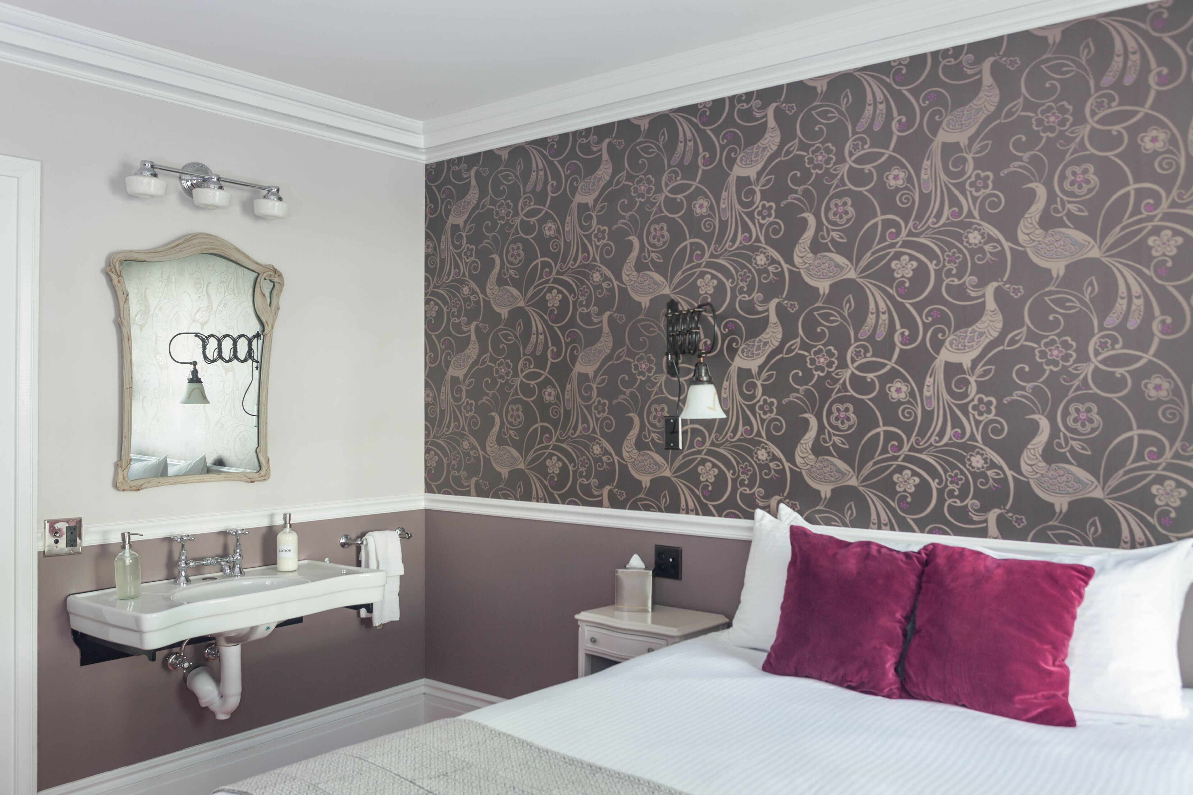 A hotel room with a large bed, a vanity with a mirror, a nightstand, and a wall with floral wallpaper.