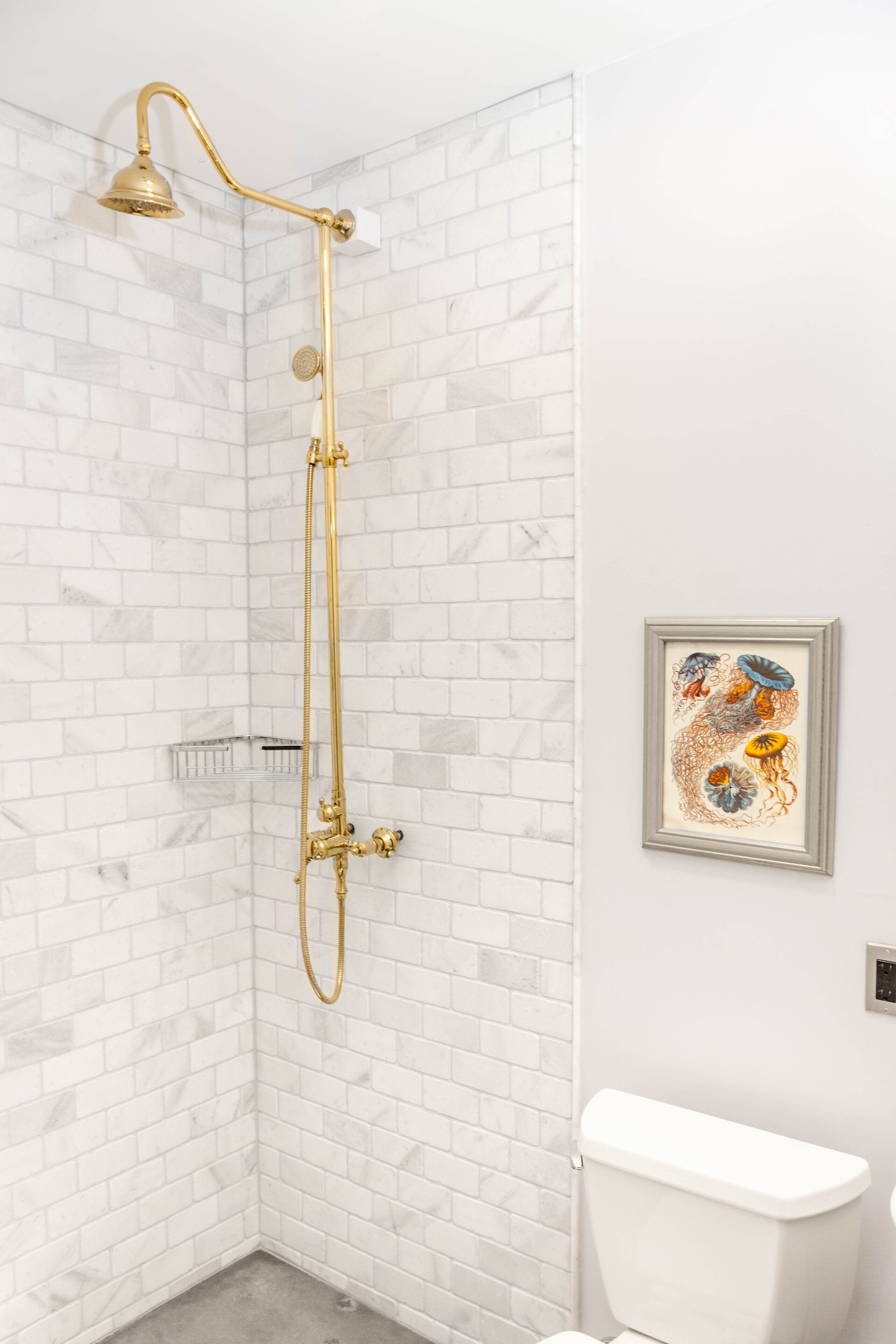 A corner shower with a gold showerhead and hose, white marble tiles, and a framed artwork on the wall.
