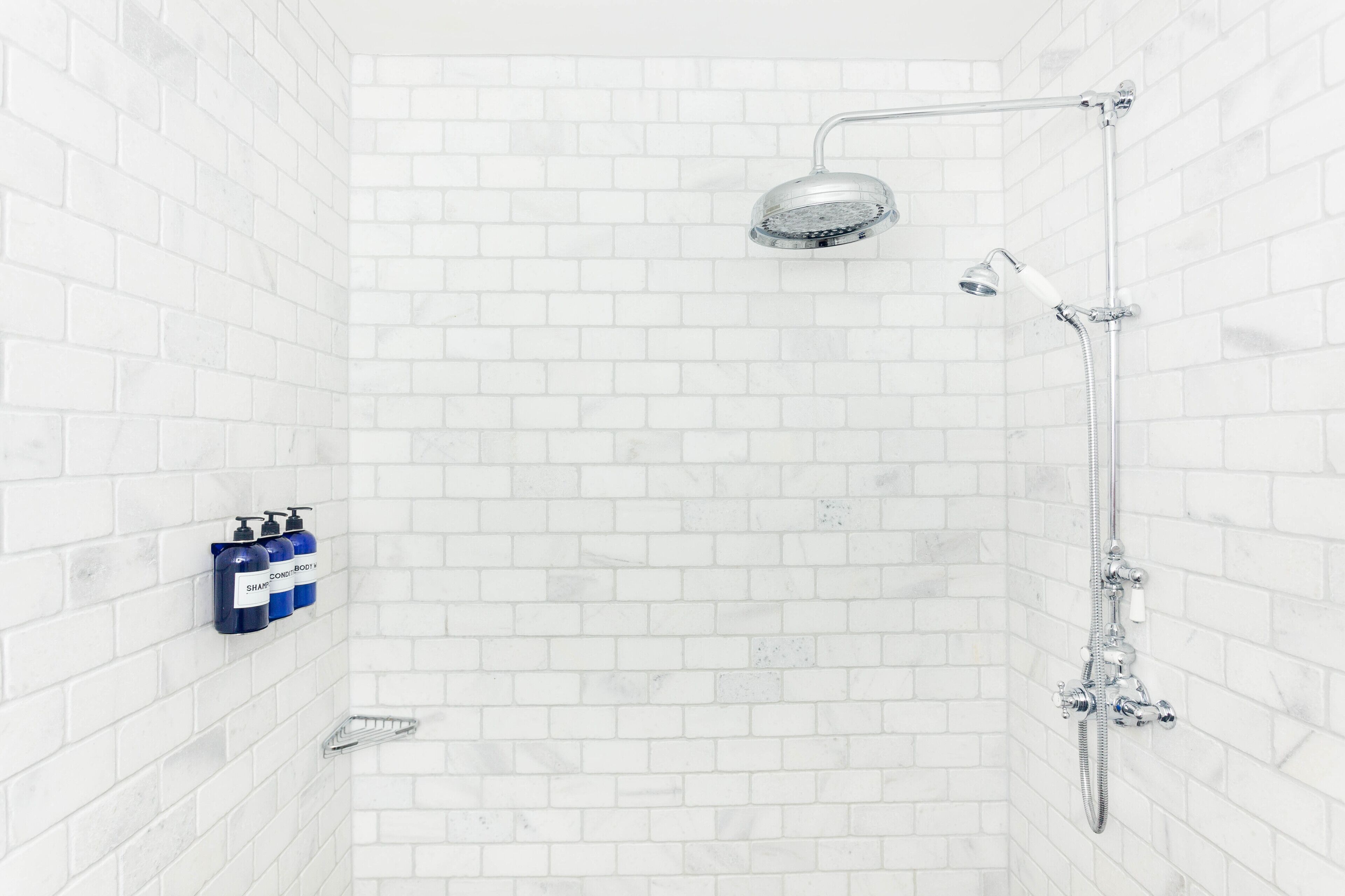 Ducha con cabezal de ducha manual y grifo empotrado en la pared. Se observan tres botellas de champú y acondicionador en la pared.