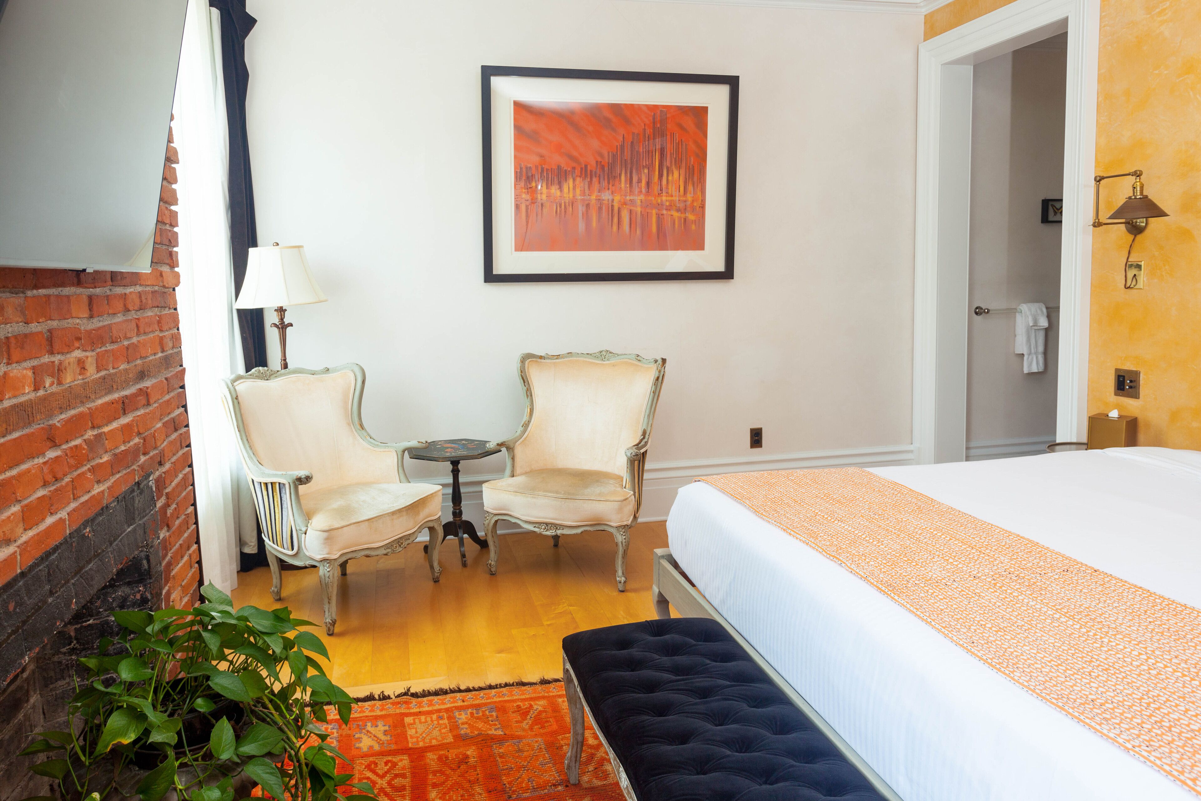 A hotel room with a bed, two chairs, a small table, a lamp, a fireplace, and a painting on the wall.