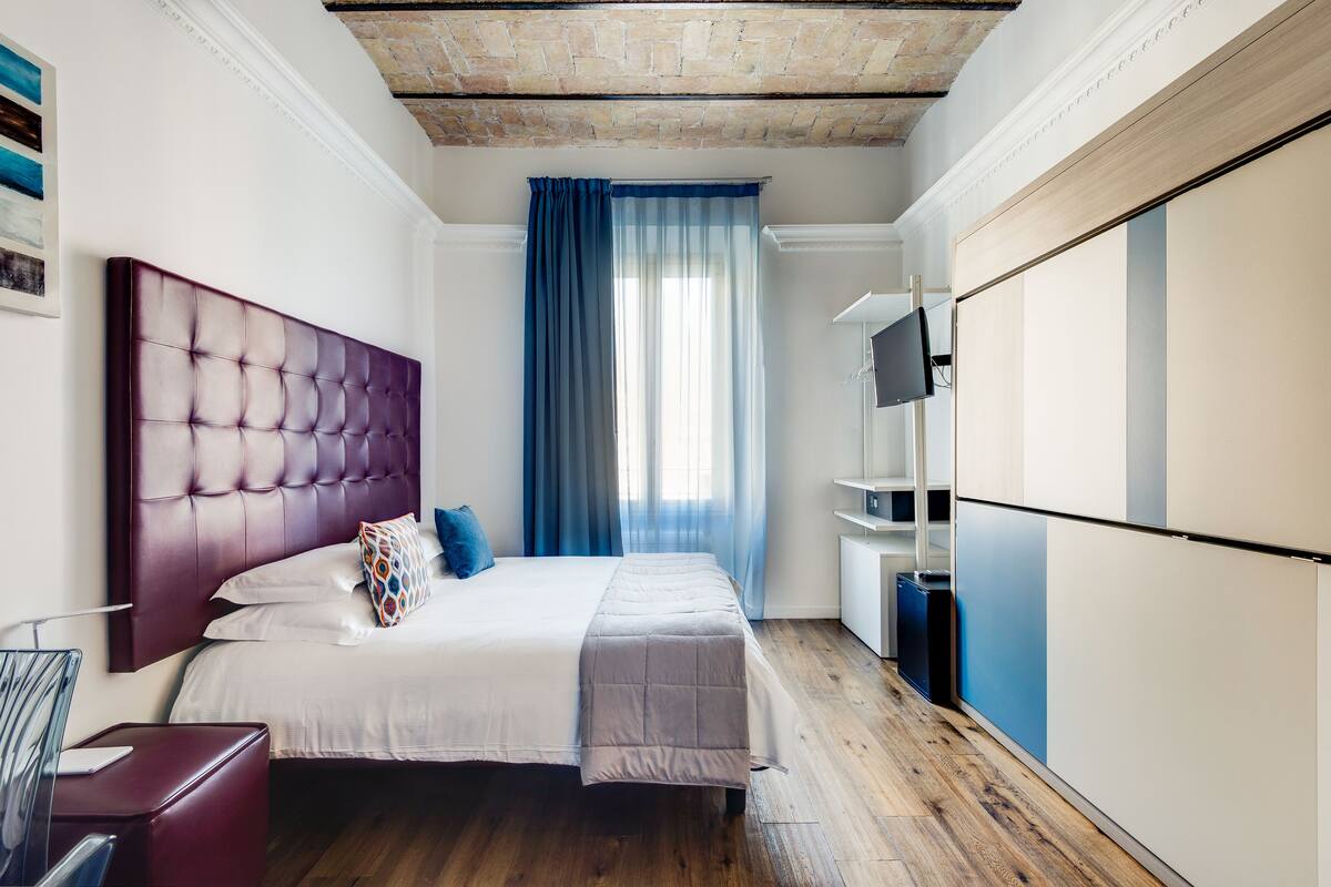A modern bedroom with a tufted headboard, a large bed, a wardrobe, and a window with blue curtains.