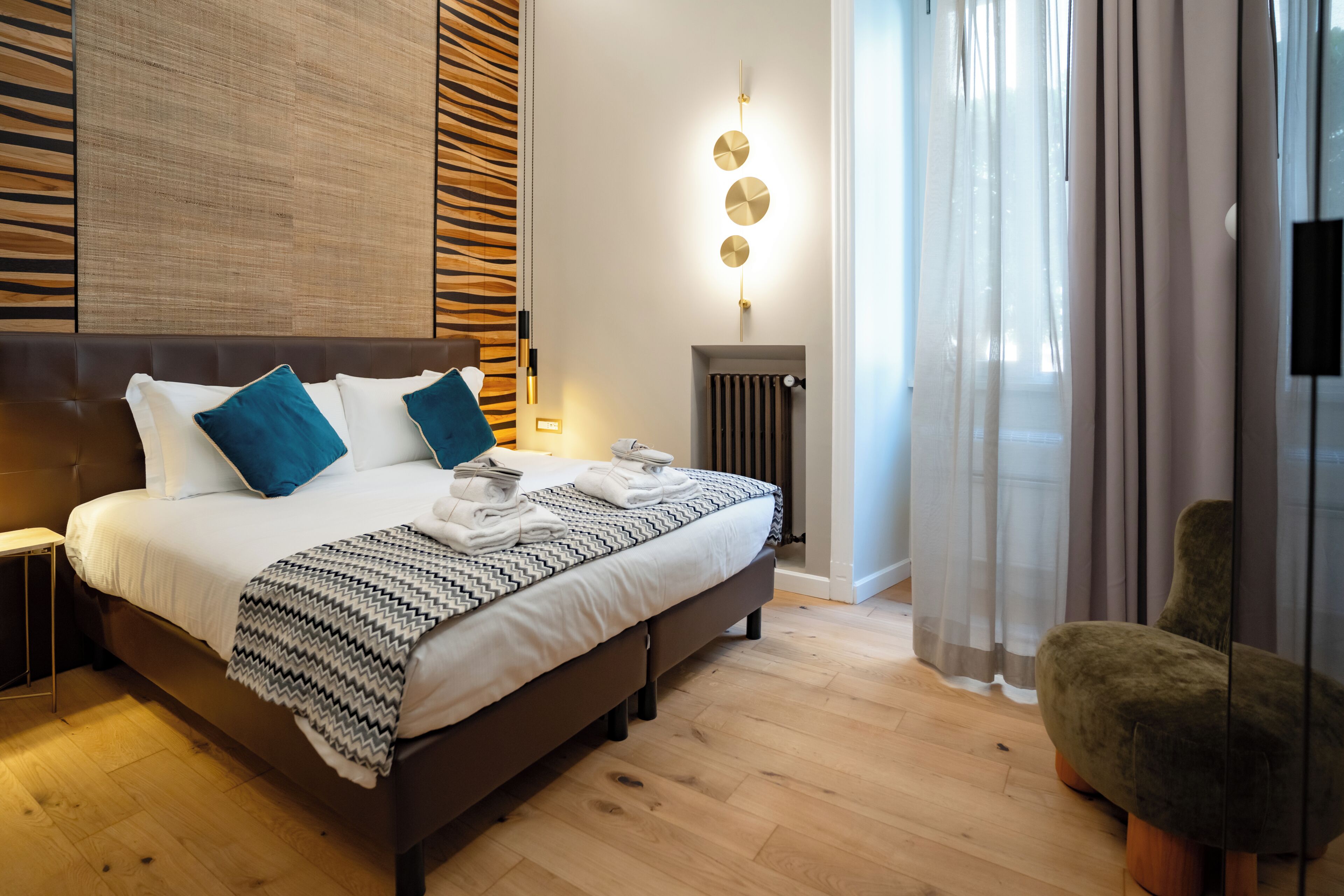 A modern hotel room with a large bed, a headboard with a textured pattern, a bedside table, a wall-mounted light fixture, a radiator, and a view of the outdoors through sheer curtains.