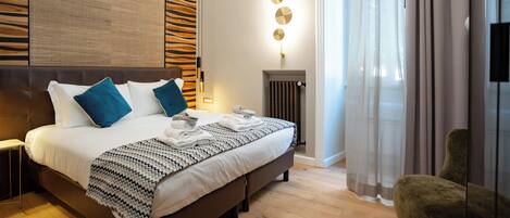 A modern hotel room with a large bed, a headboard with a textured pattern, a bedside table, a wall-mounted light fixture, a radiator, and a view of the outdoors through sheer curtains.