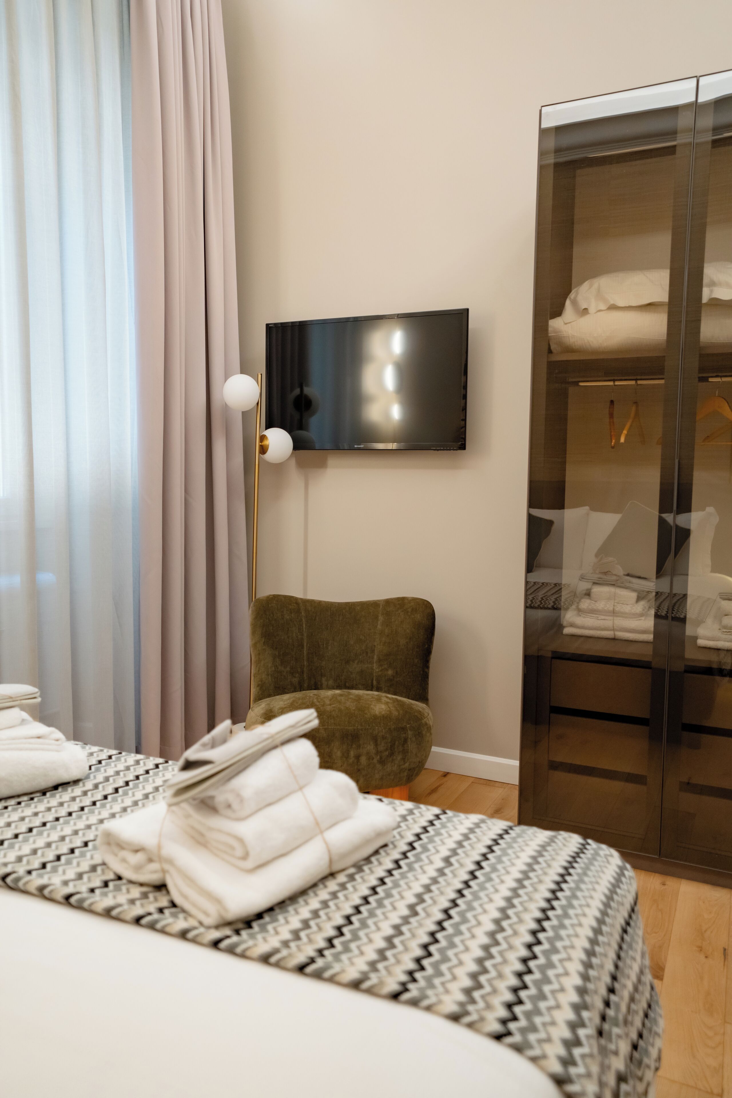 A hotel room with a bed, a flat-screen TV mounted on the wall, a wardrobe, and a floor lamp.