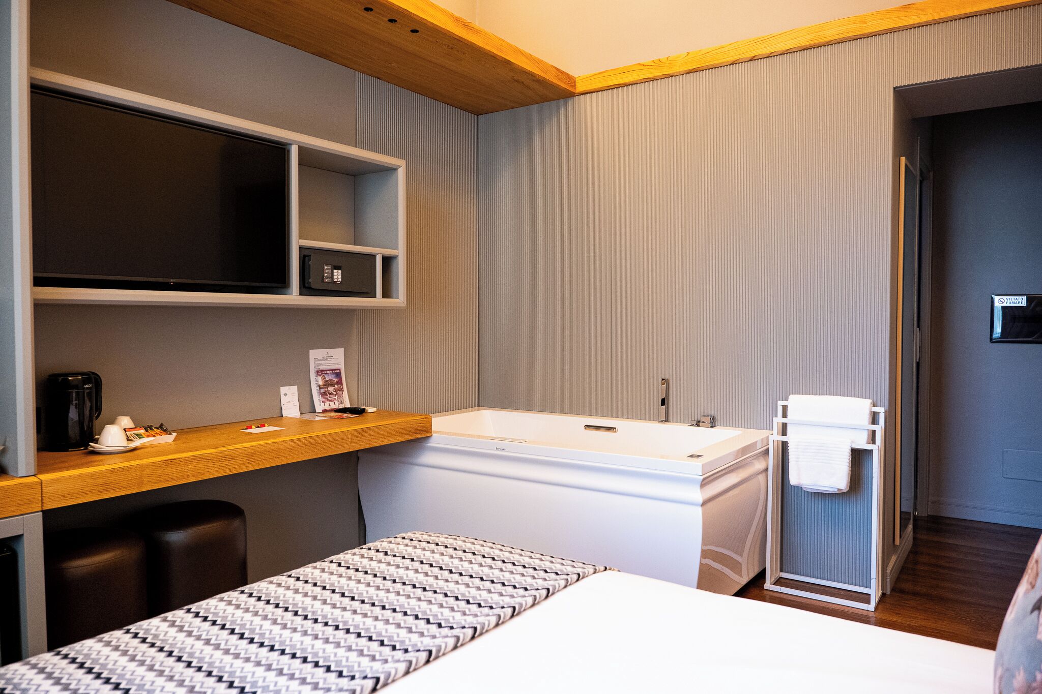 A modern hotel room with a wooden desk, a large bed, a built-in TV, a bathtub, and a shower area.
