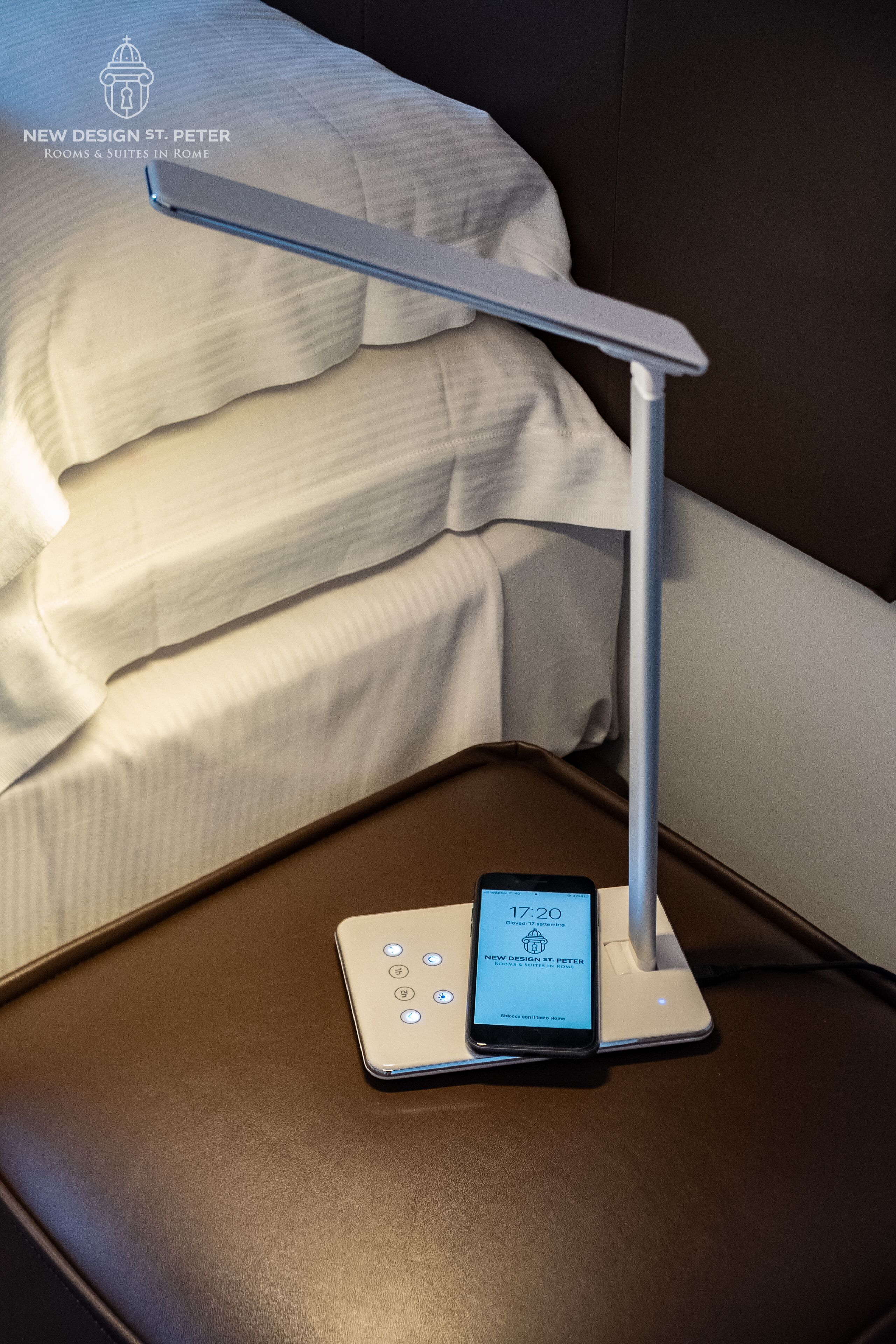 Uma cama com edredom branco e travesseiros, uma mesa de cabeceira com um smartphone carregando e um abajur branco.