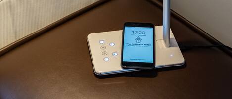 Uma cama com edredom branco e travesseiros, uma mesa de cabeceira com um smartphone carregando e um abajur branco.
