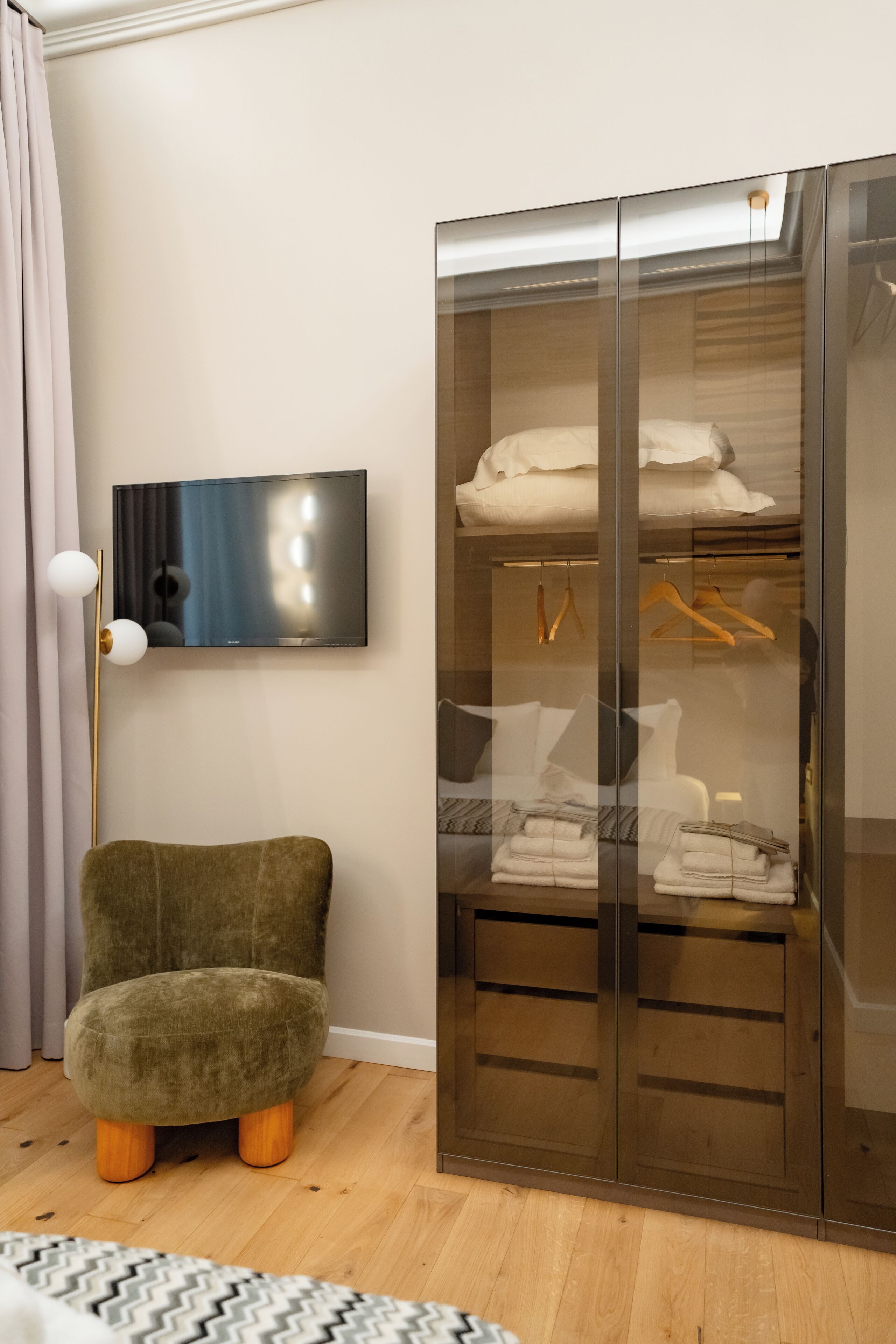 A room with a green armchair, a wardrobe with glass doors, and a flat-screen TV mounted on the wall.
