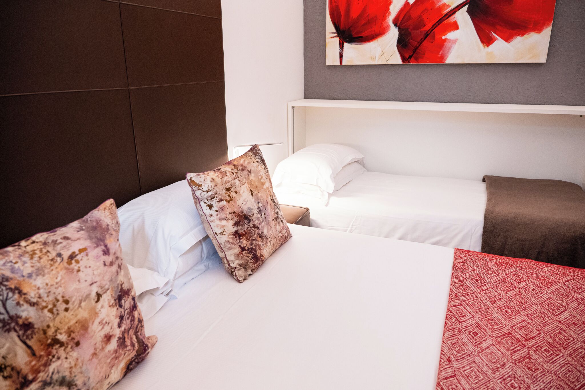 A hotel room with a bed, pillows, a red patterned blanket, and a painting on the wall.