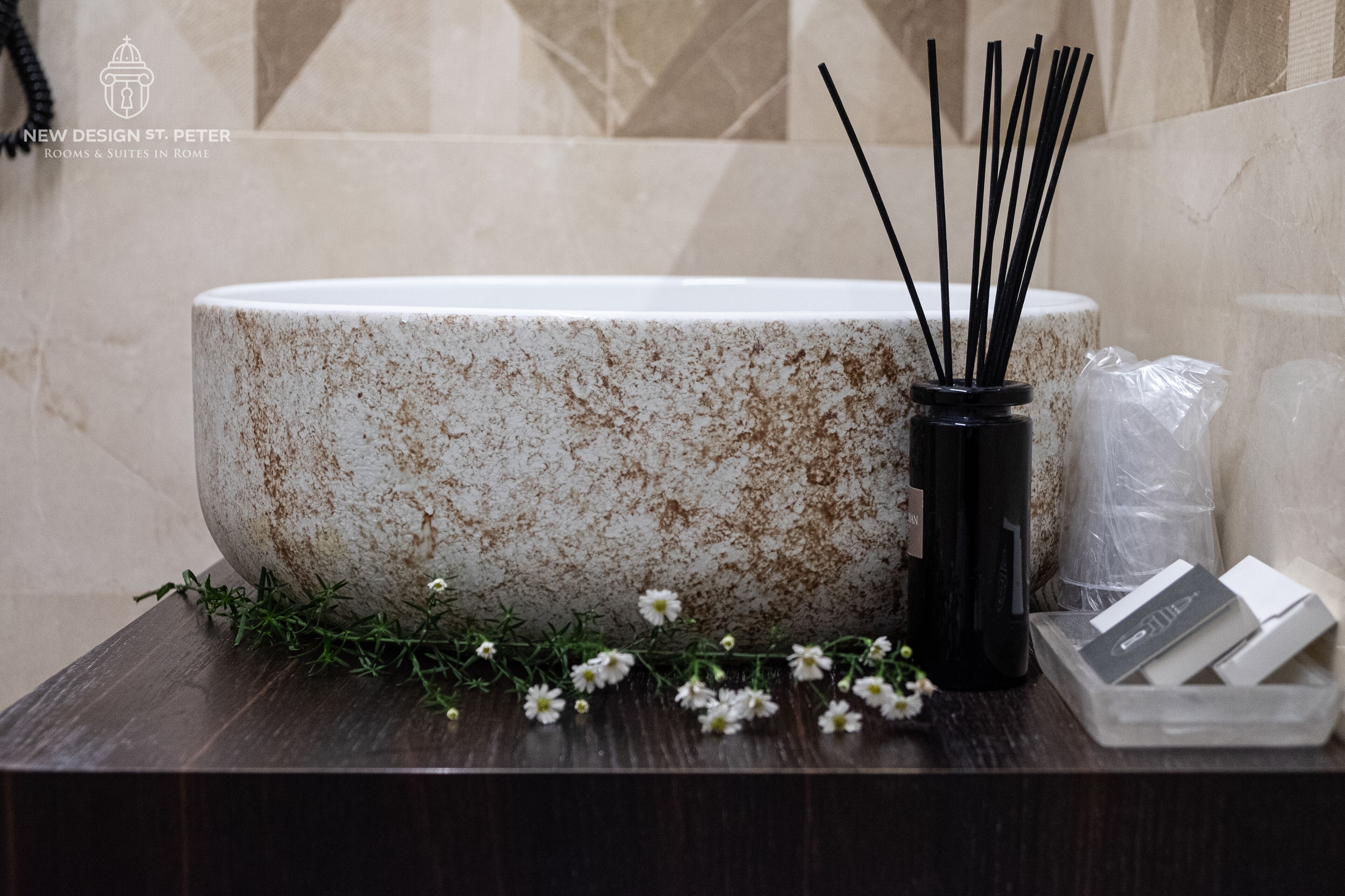 A bathroom sink with a speckled design, a black vase with reed sticks, and a sprig of greenery.