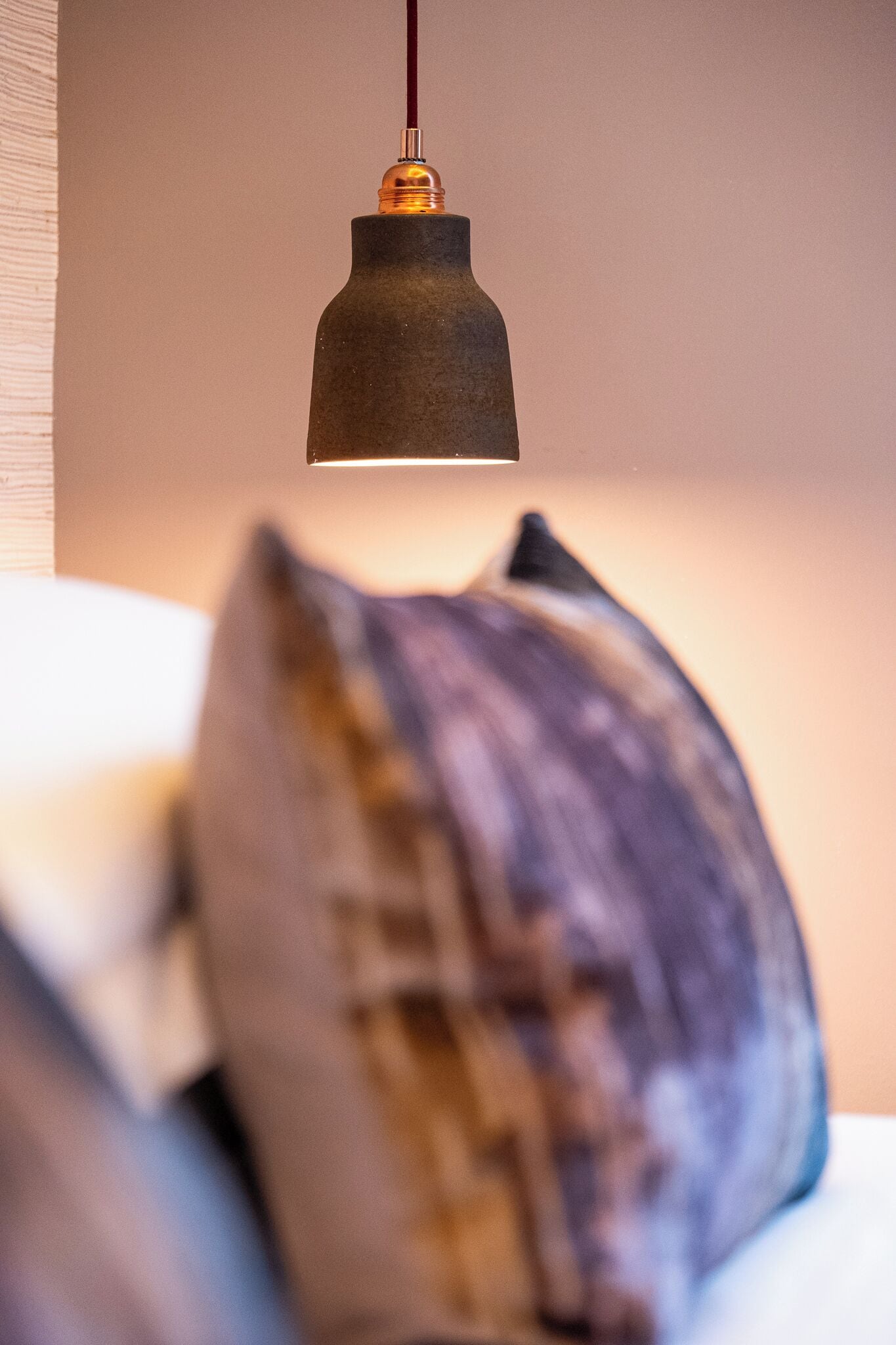 Un luminaire suspendu, de type suspension, avec un abat-jour foncé et un dessus de couleur cuivrée, placé au-dessus d’un objet flou.