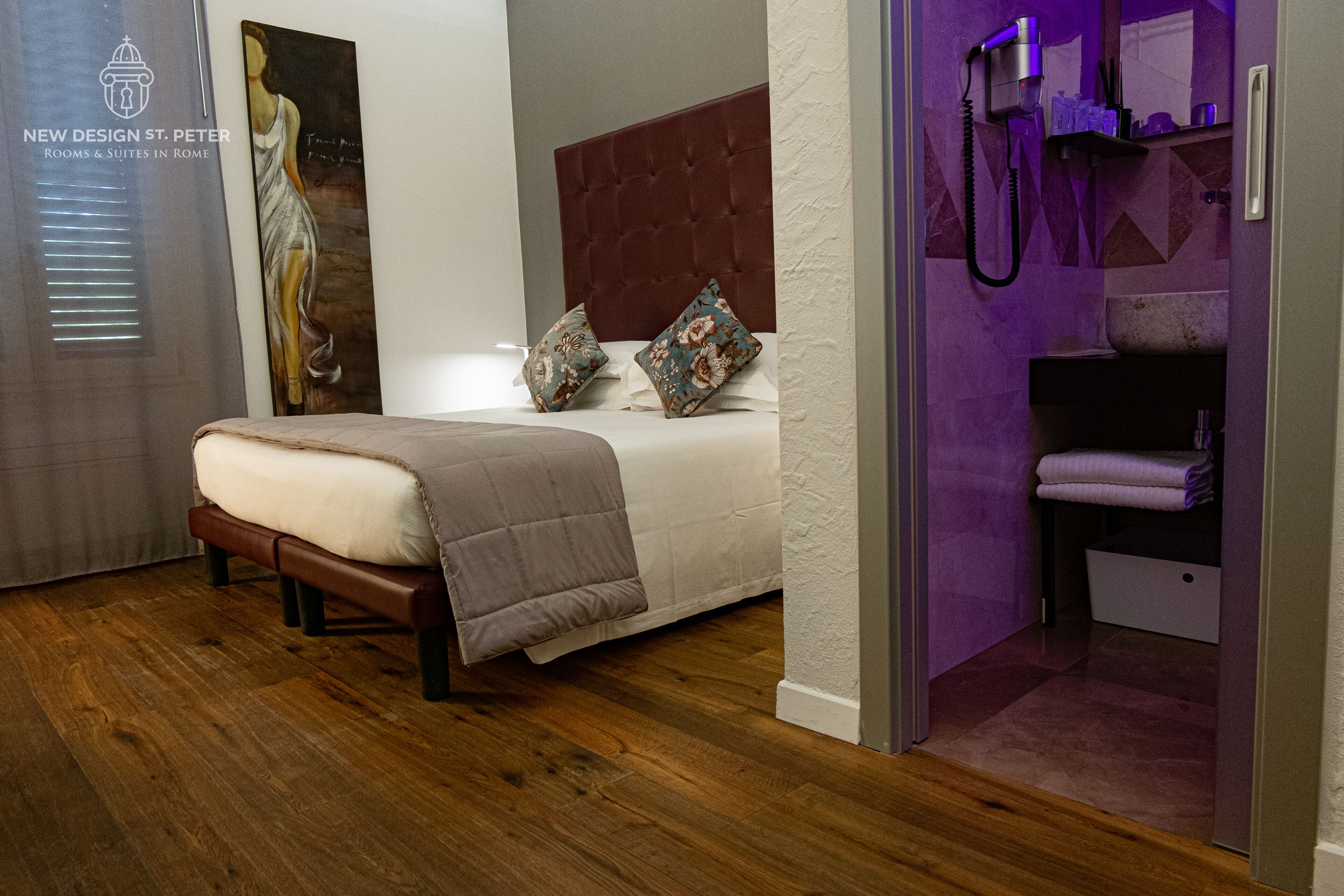 A modern bedroom with a wooden floor, a bed with a brown headboard, two decorative pillows, and a painting on the wall.