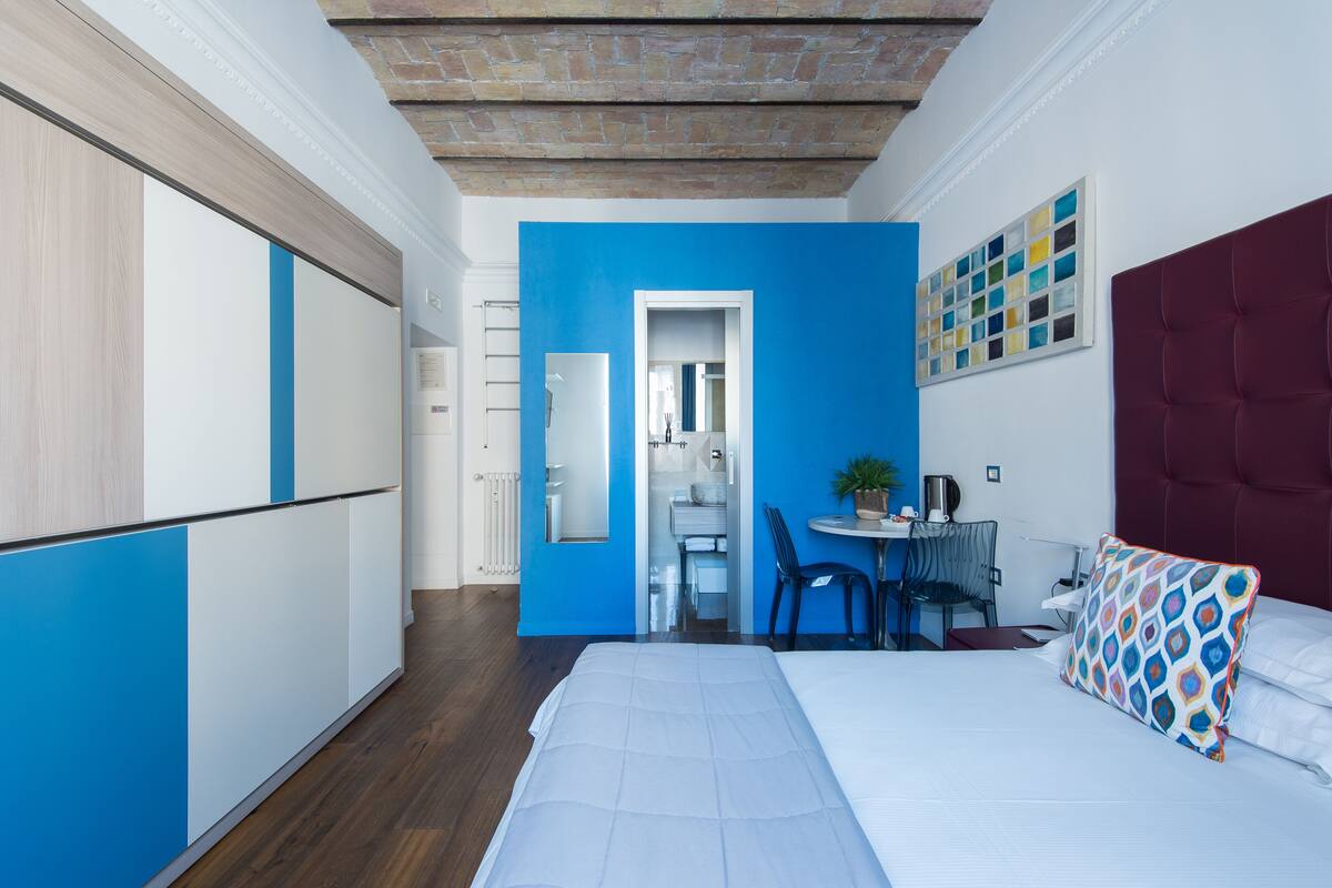 A modern hotel room with a bed, a desk, and a chair. There is a bathroom visible through an open door.