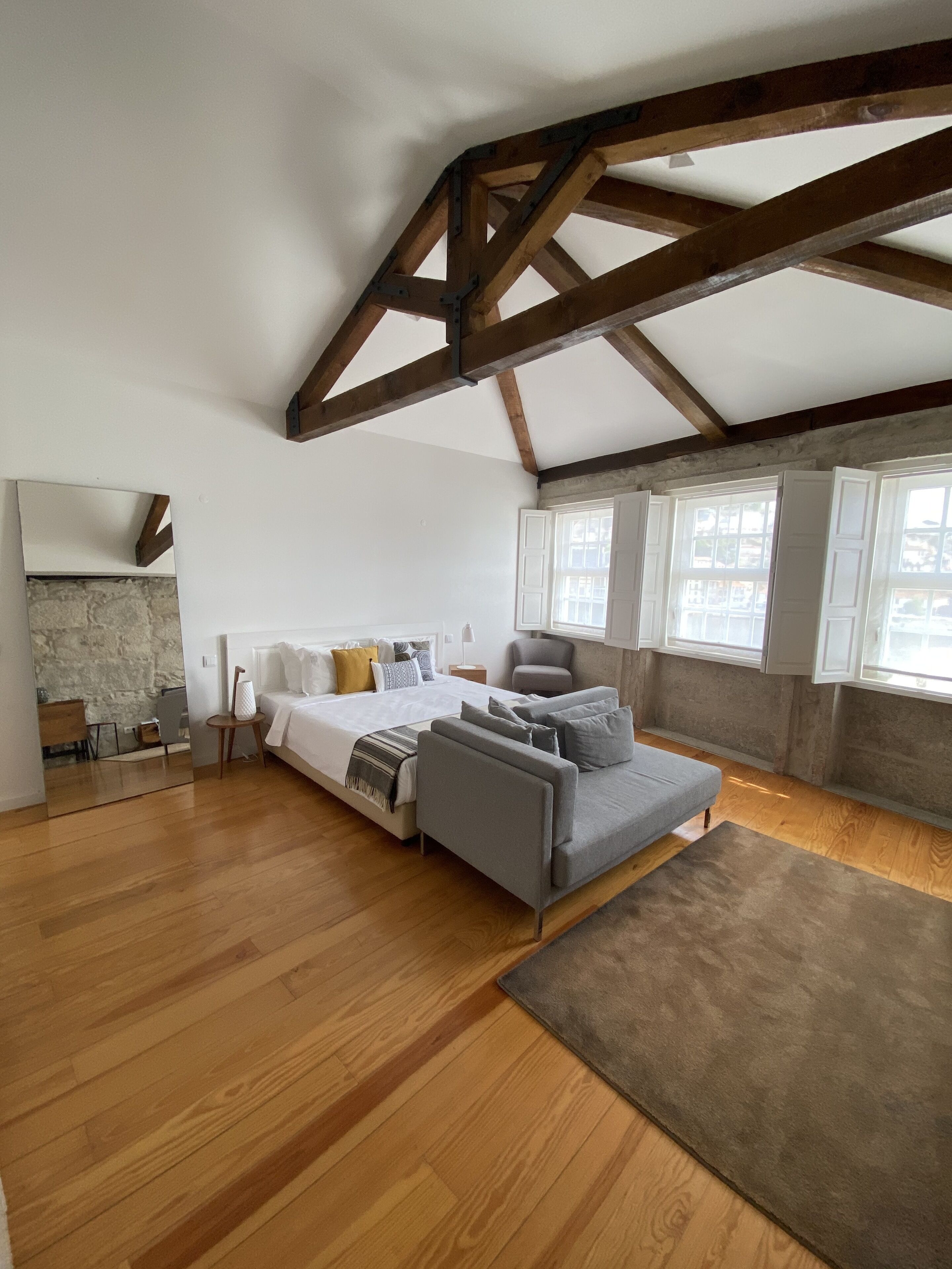 Una habitación amplia con una cama grande, un sofá gris, suelo de madera y una chimenea de piedra.