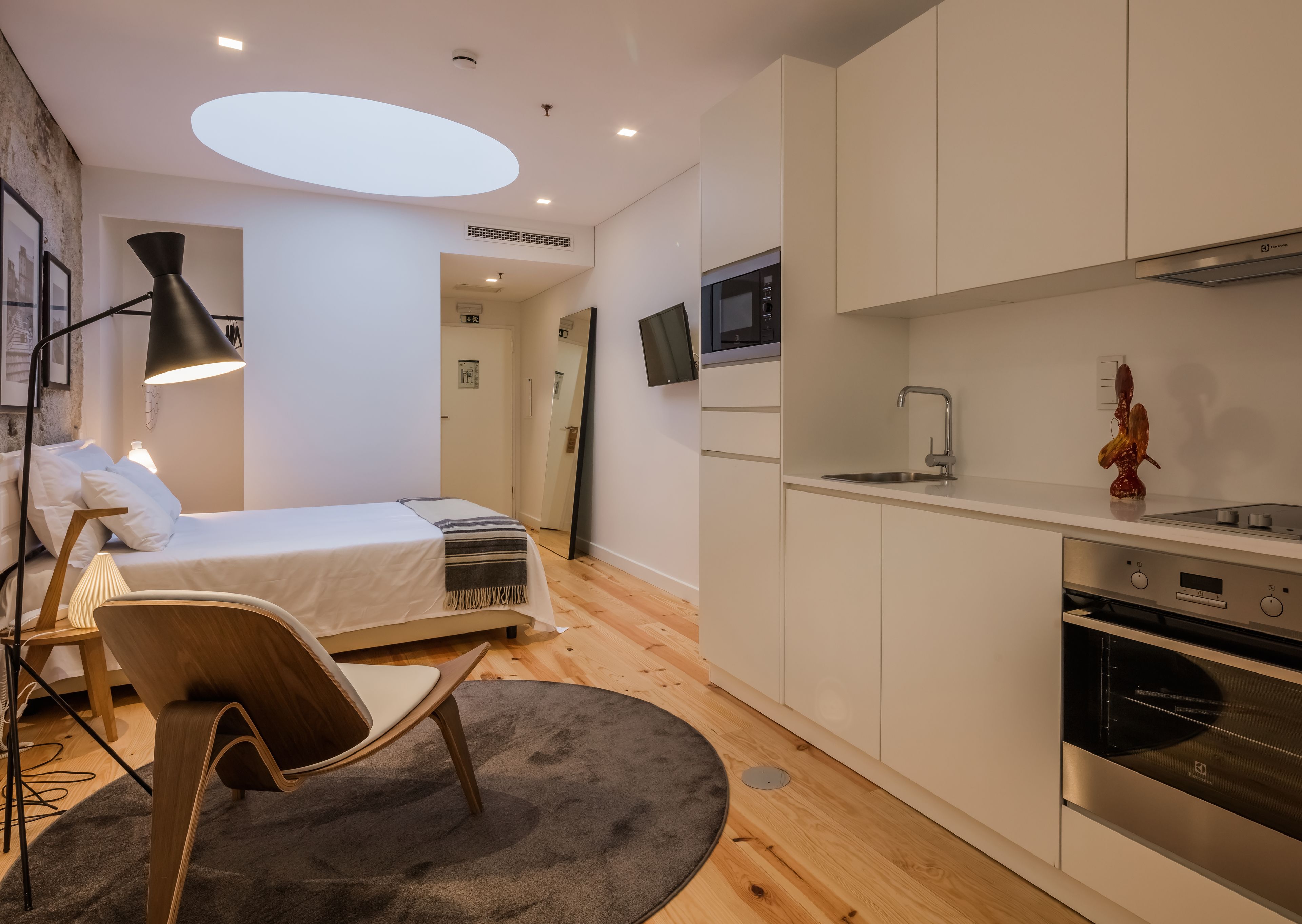 A modern bedroom with a bed, bedside lamp, and a chair. There is a kitchenette with a microwave, sink, and oven.