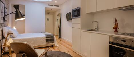 A modern bedroom with a bed, bedside lamp, and a chair. There is a kitchenette with a microwave, sink, and oven.