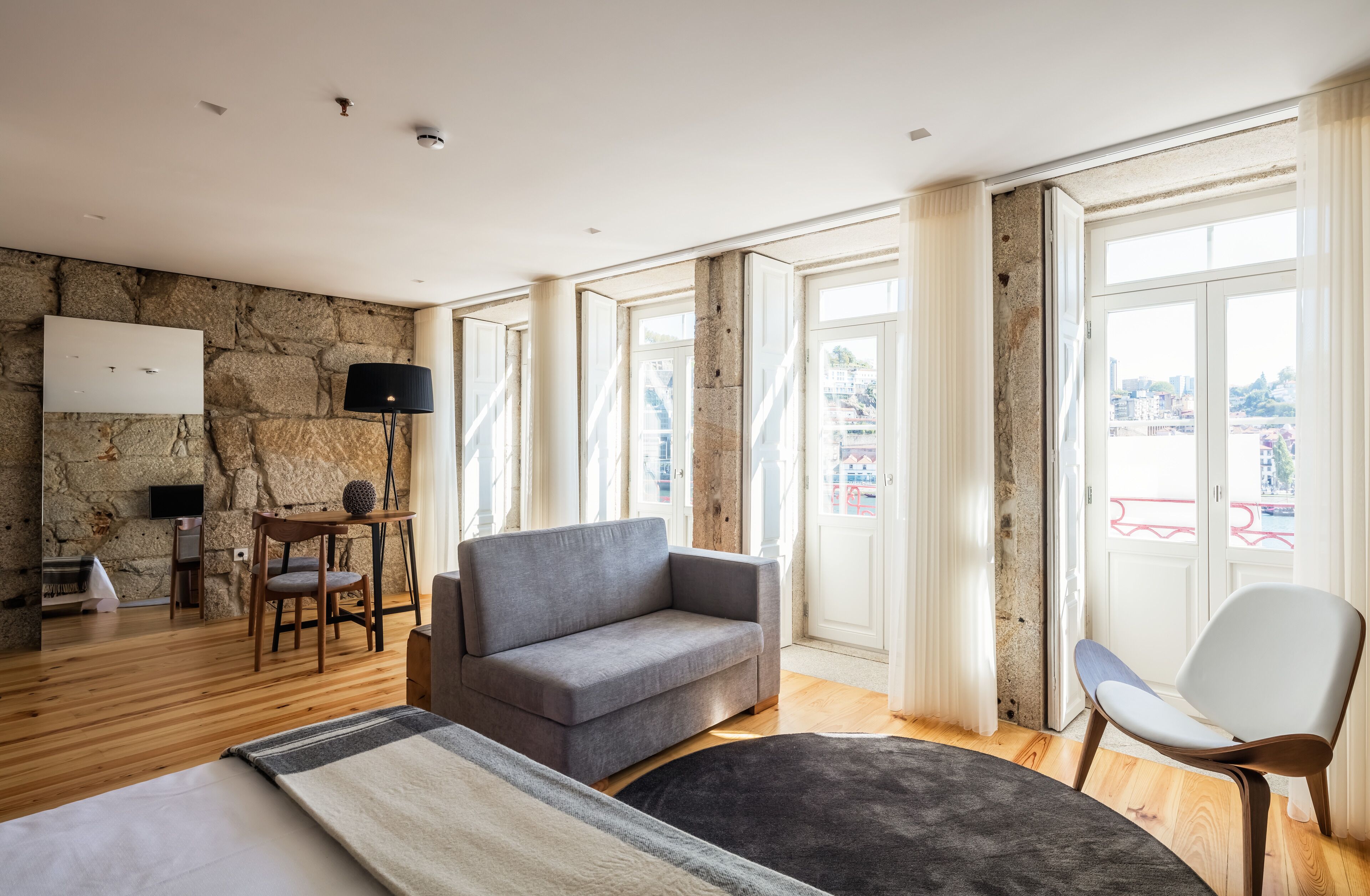 Habitación de hotel moderna con pared de piedra, sofá gris, silla blanca, suelo de madera y ventanas grandes con cortinas.