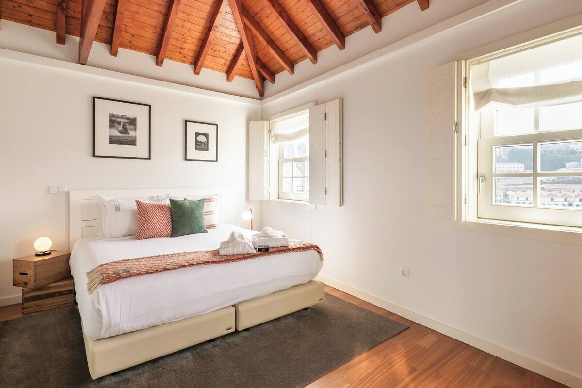 Une chambre avec un plafond en bois, un lit recouvert de linge de lit blanc, deux tableaux encadrés au mur et une fenêtre donnant sur un paysage.