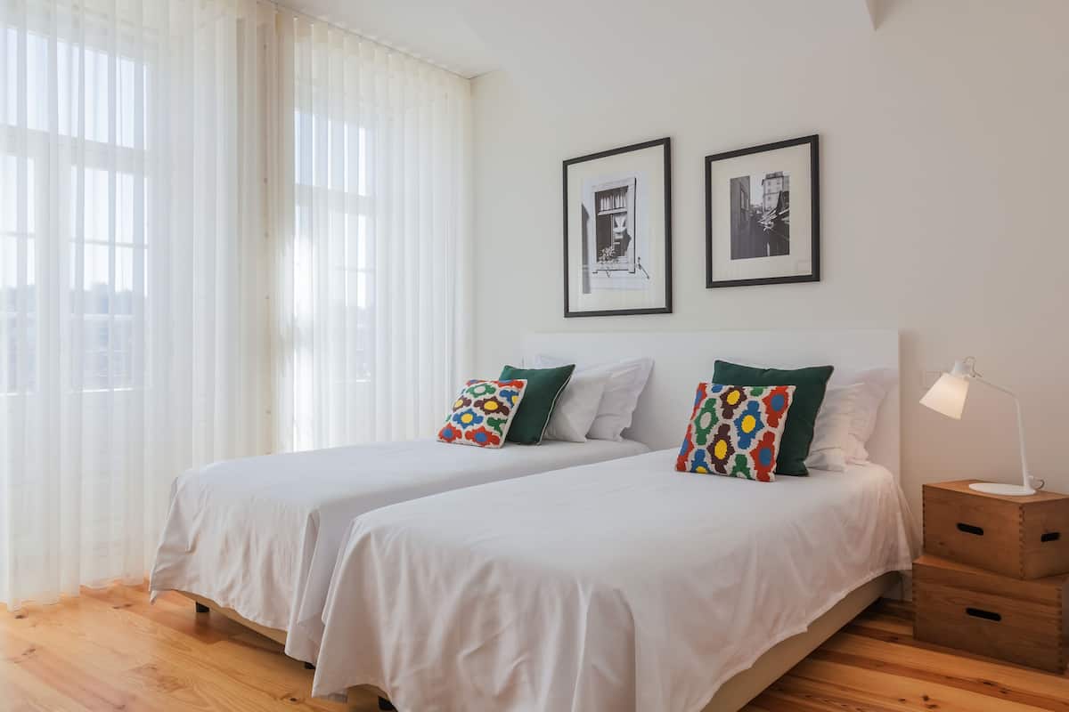 Une chambre avec deux lits, du linge de lit blanc, des oreillers verts et des tables de chevet en bois. Deux tableaux encadrés ornent le mur, et une fenêtre est agrémentée de rideaux légers.