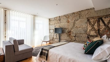 A modern bedroom with a stone wall, a bed with white linens, a grey sofa, and a wooden floor.