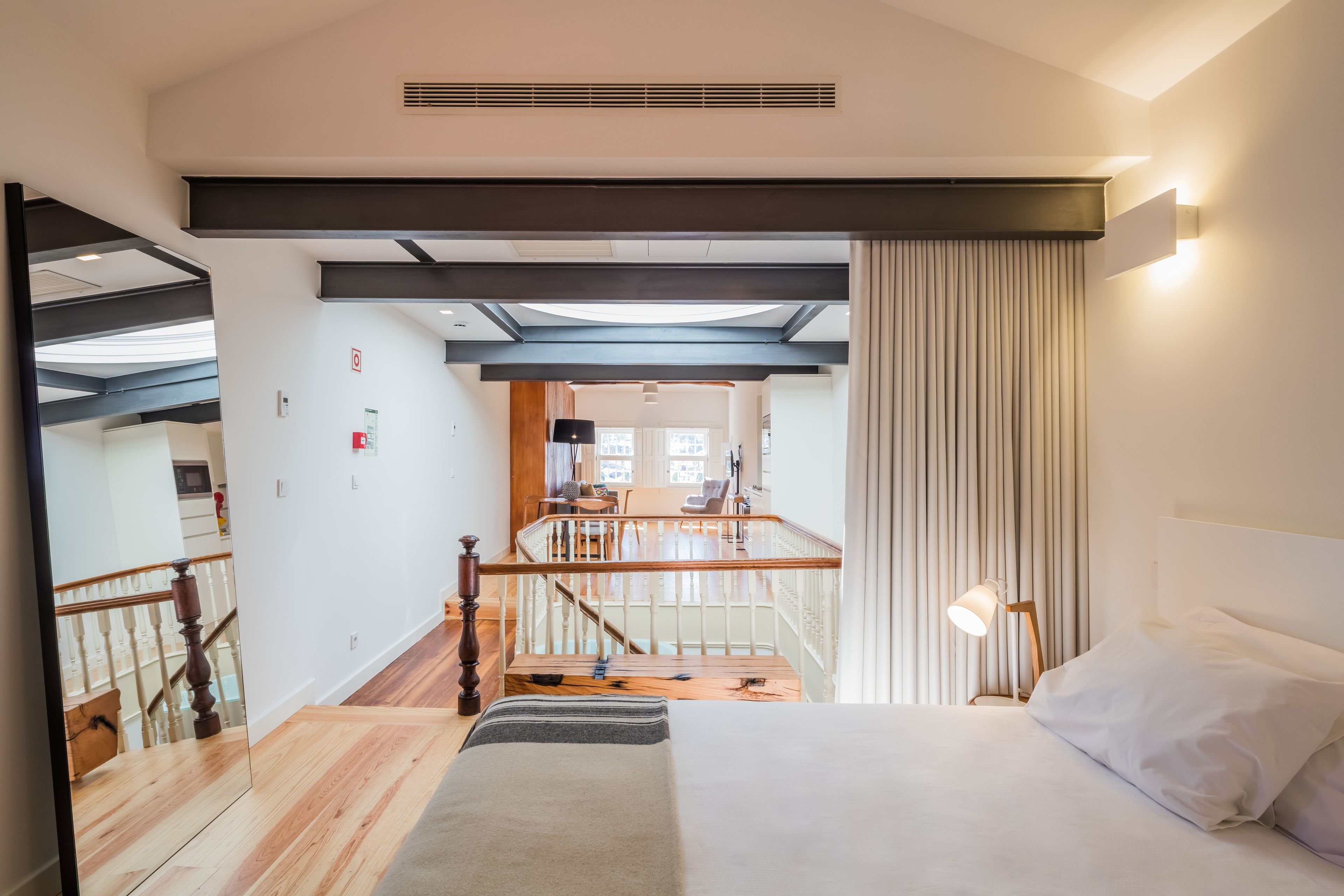 A modern hotel room with a large bed, a wooden staircase, a balcony, and a view of the dining area.