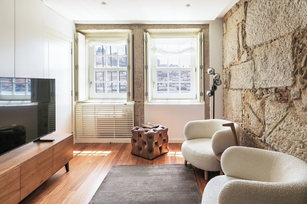 Un salon moderne doté d’un grand téléviseur à écran plat, de deux fenêtres avec des stores blancs, d’un mur en pierre et d’un canapé beige.