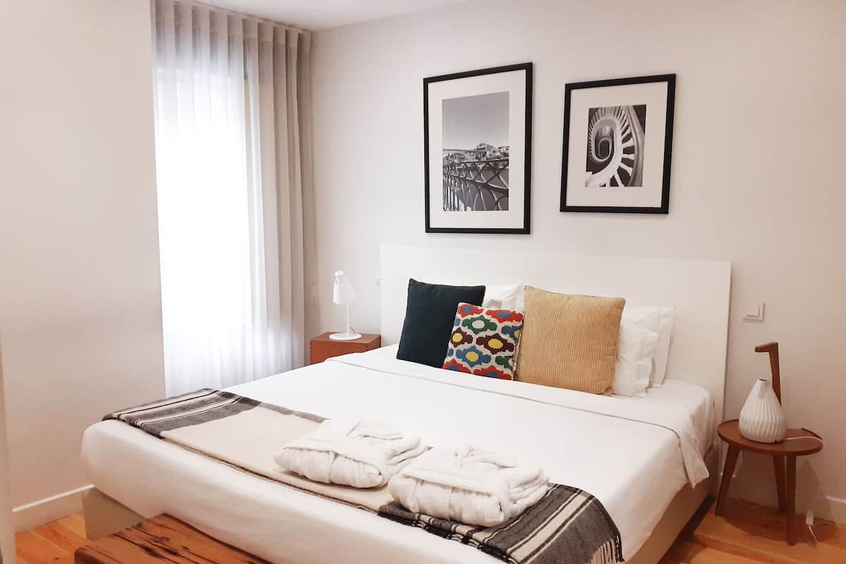 Un lit bien fait, avec une couette blanche et deux oreillers, une table de chevet avec une lampe, et deux tableaux encadrés accrochés au mur.