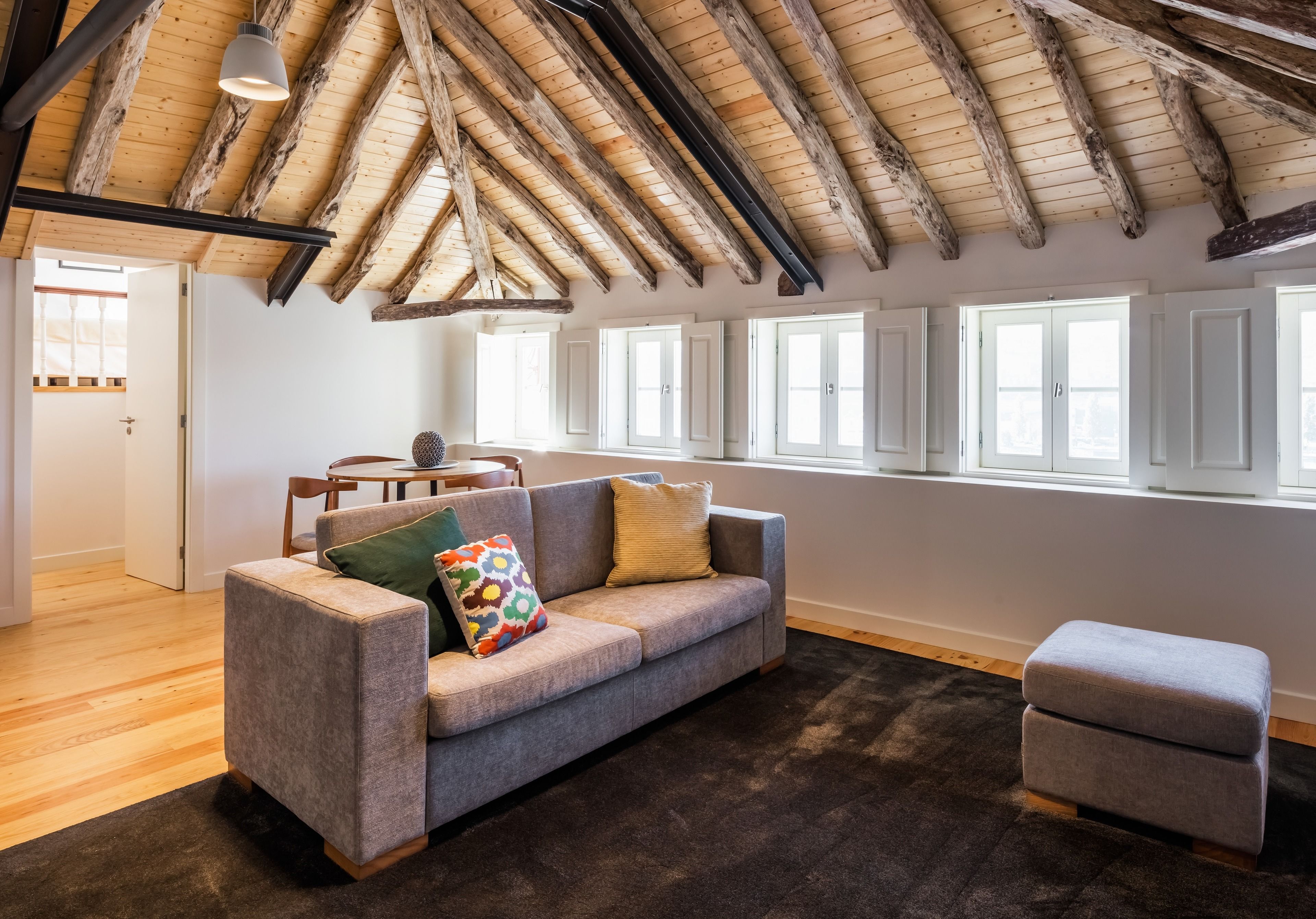 A spacious living room with a high wooden ceiling, a grey sofa with colorful pillows, a small ottoman, and large windows allowing natural light.