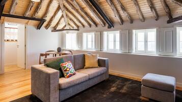 A spacious living room with a high wooden ceiling, a grey sofa with colorful pillows, a small ottoman, and large windows allowing natural light.