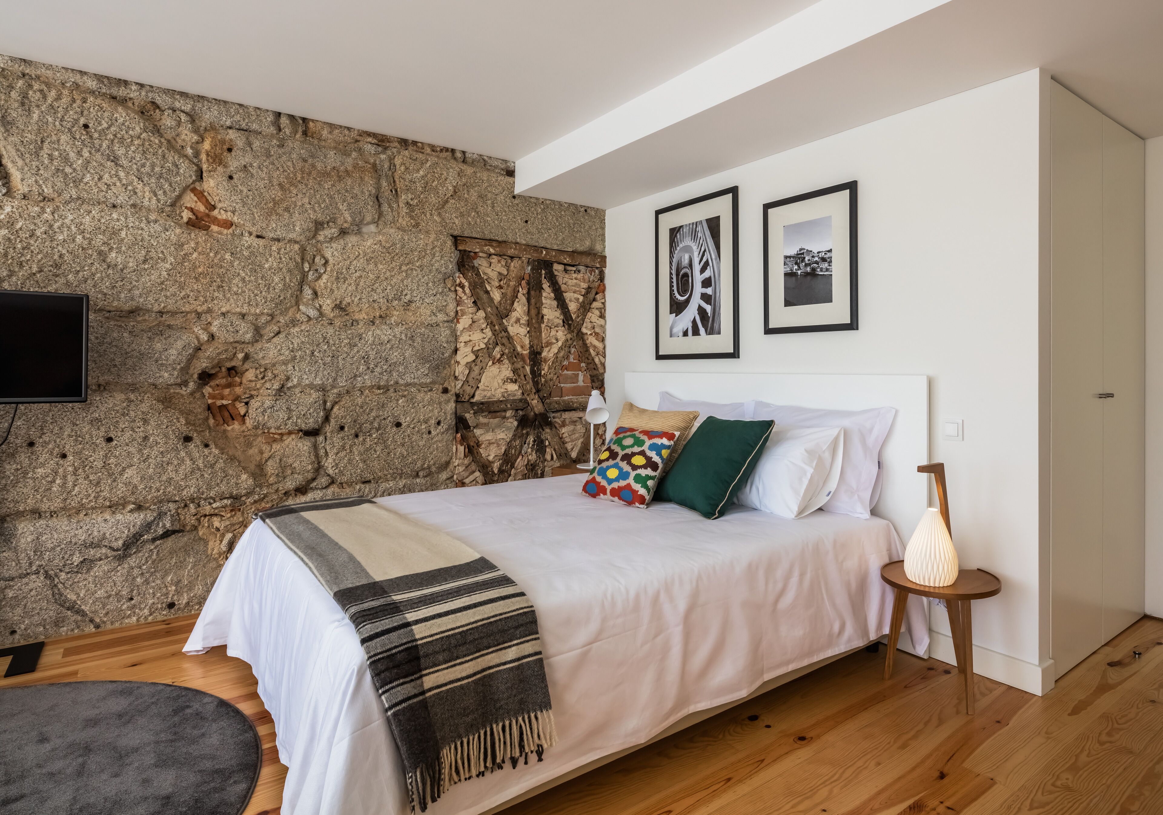 A bedroom with a stone wall, a bed with pillows, a bedside table with a lamp, and two framed pictures on the wall.