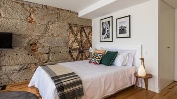 A bedroom with a stone wall, a bed with pillows, a bedside table with a lamp, and two framed pictures on the wall.