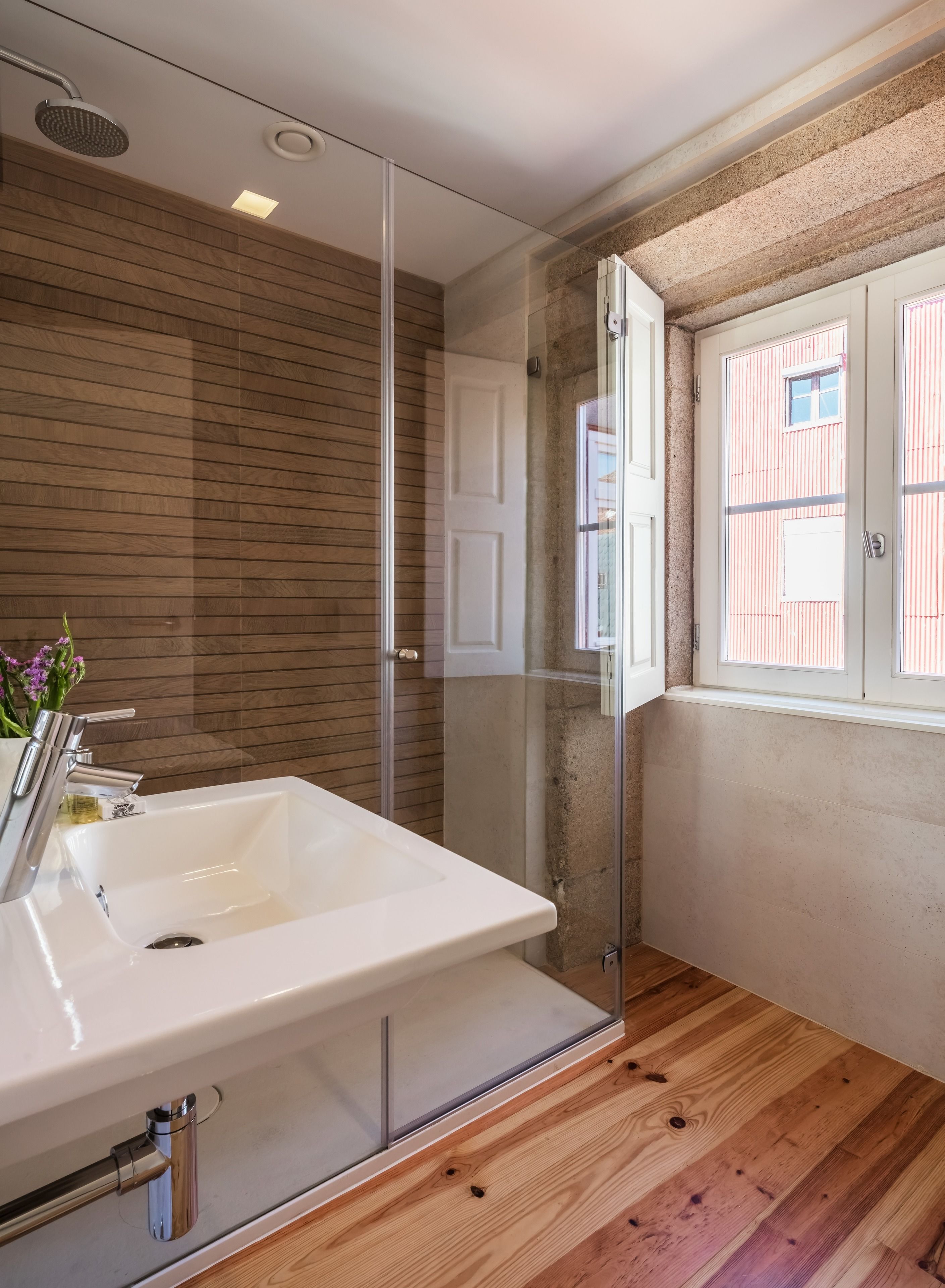 A modern bathroom with a large white sink, a glass shower enclosure, wooden flooring, and a window with striped blinds.