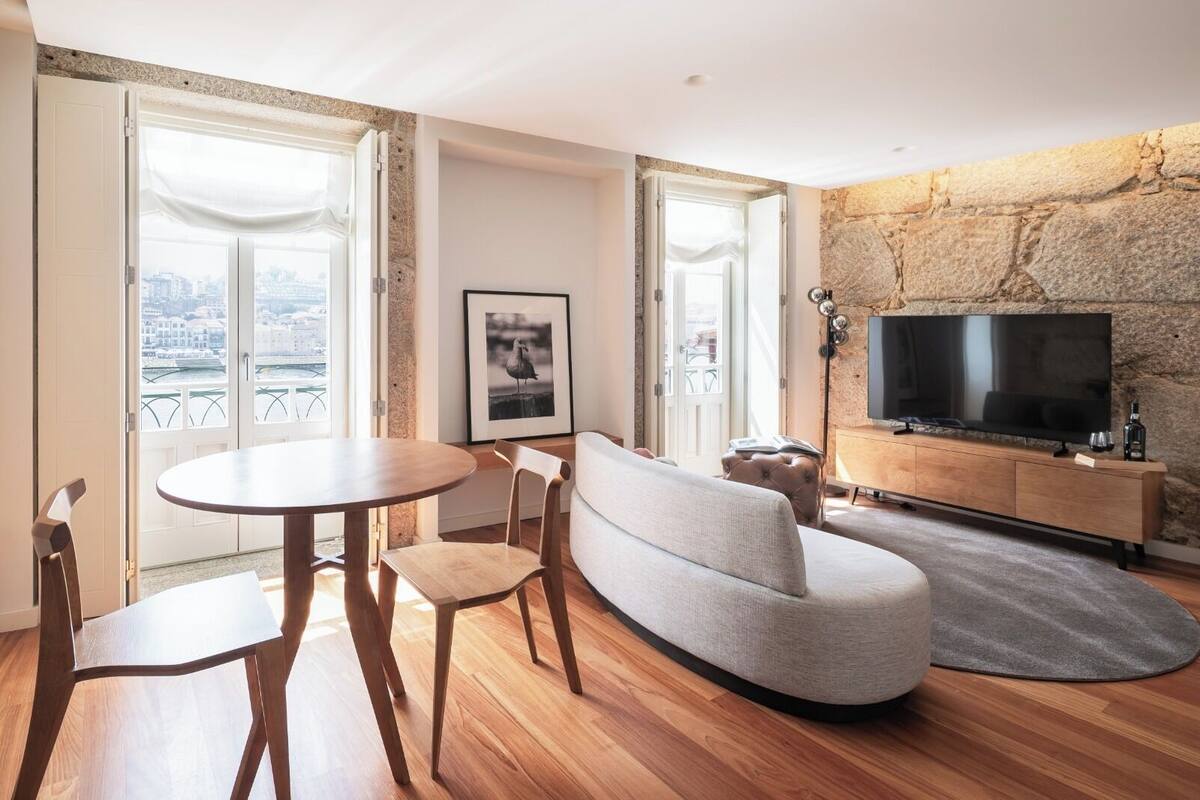 Un salon moderne avec un canapé, une table ronde et des chaises, un téléviseur fixé à un mur en pierre, et de grandes fenêtres offrant une vue dégagée.