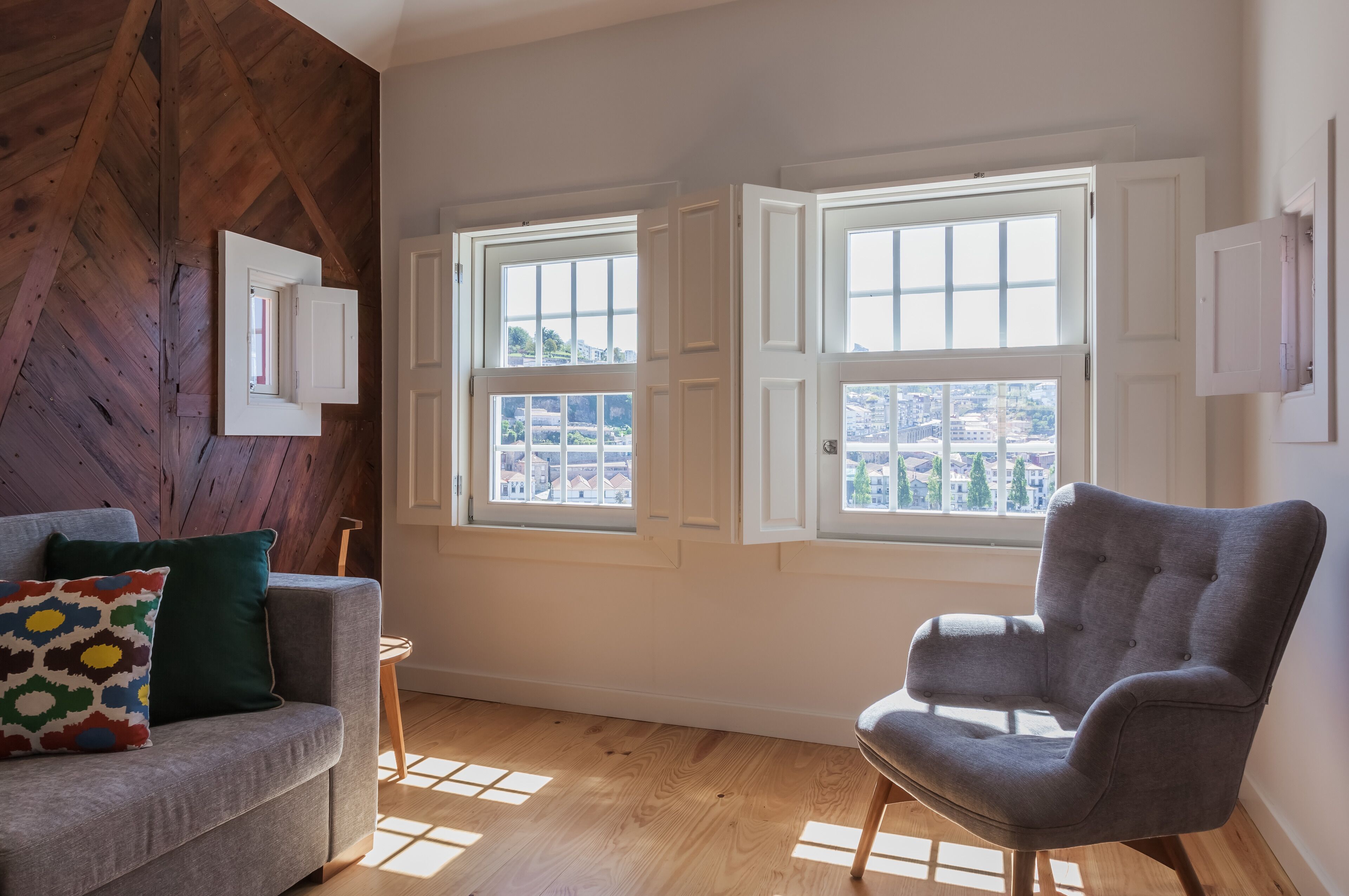 Una habitación con techo de madera, dos ventanas con contraventanas blancas, un sillón gris y un sofá gris con un cojín de colores.
