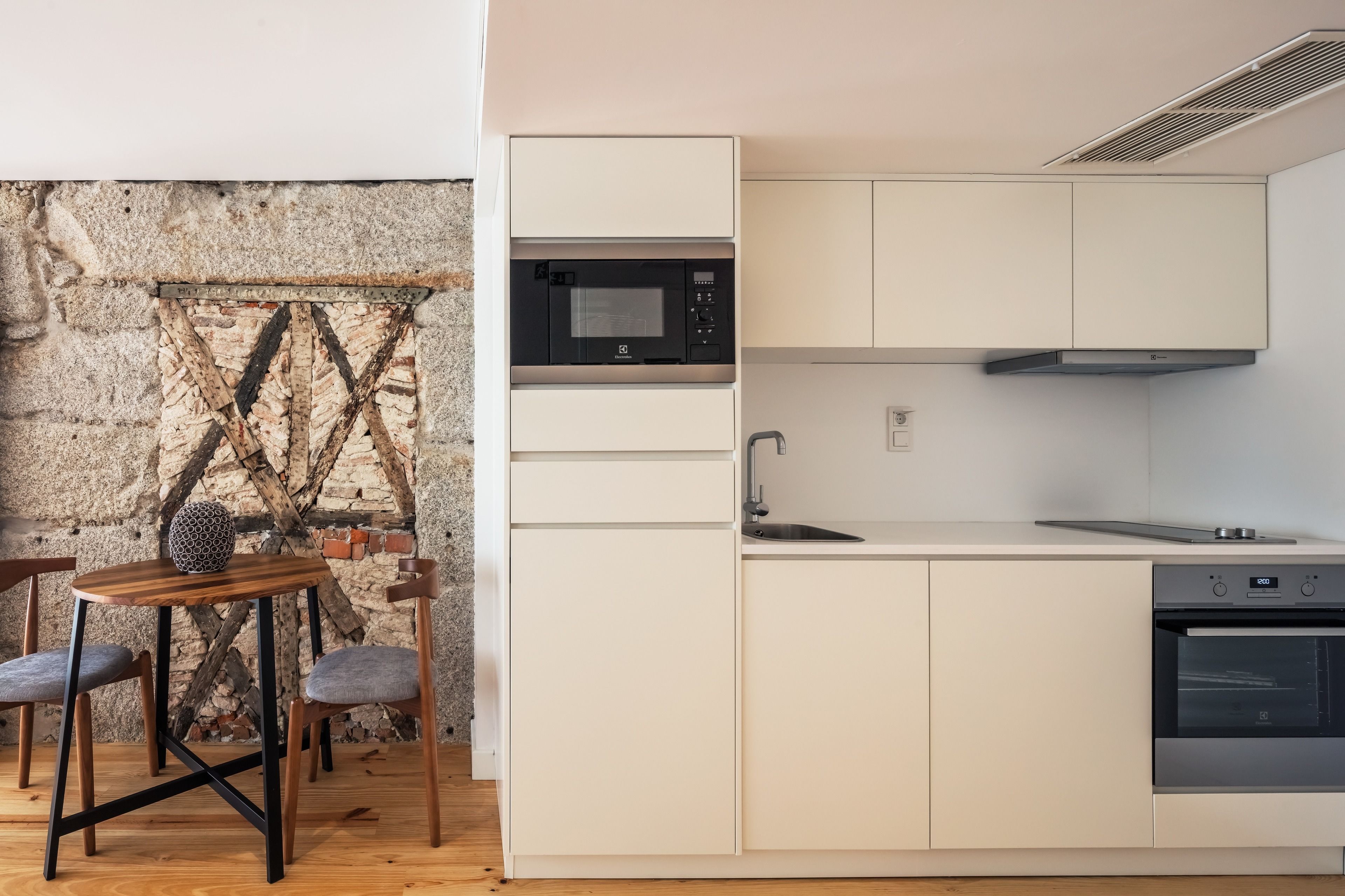Een compacte keuken met witte kastjes, een ingebouwde magnetron en een oven. Achtergrond toont een rustieke bakstenen muur en een houten vloer.