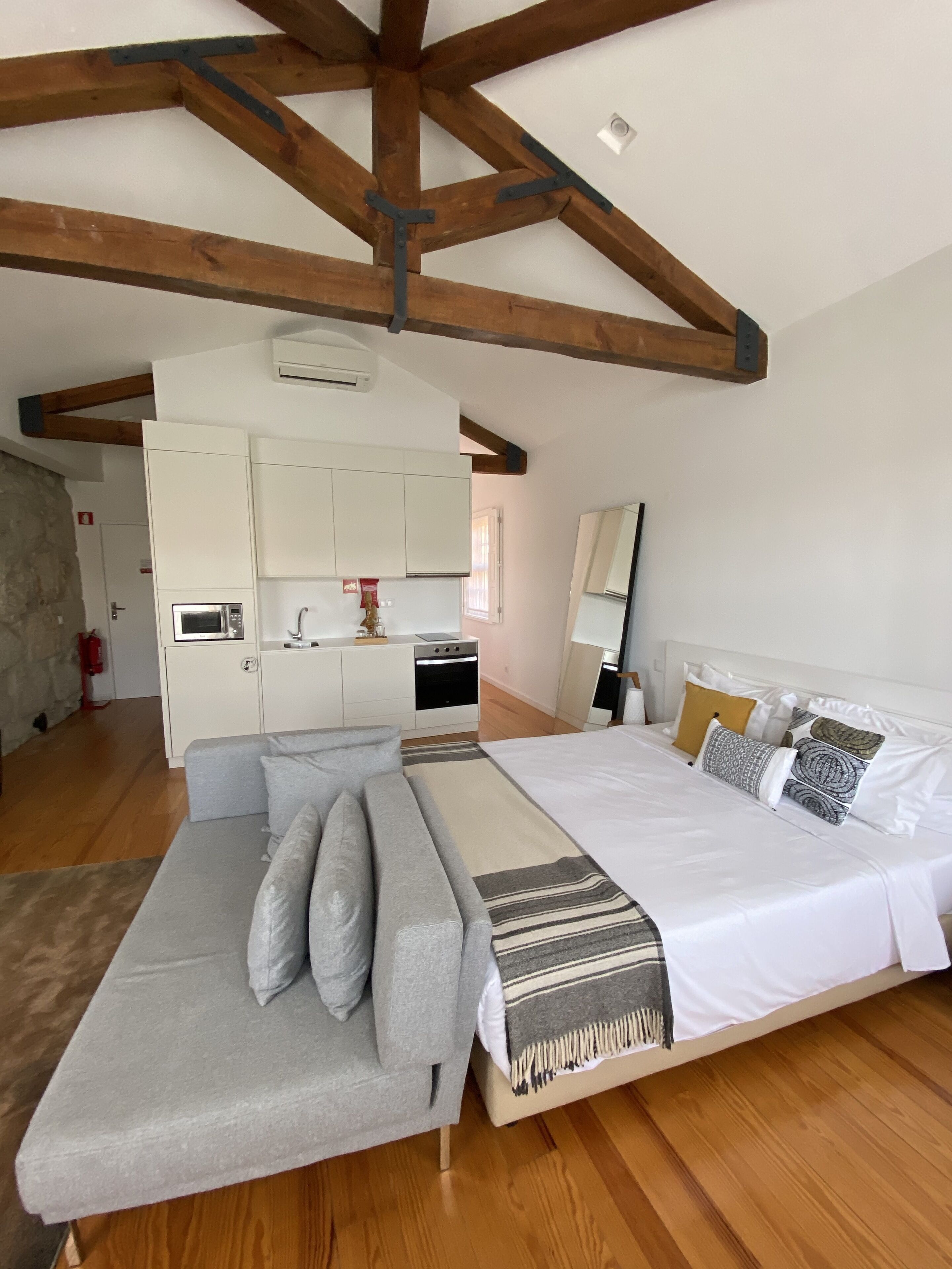 A bedroom with a bed, a sofa, a kitchen area, and wooden beams on the ceiling.