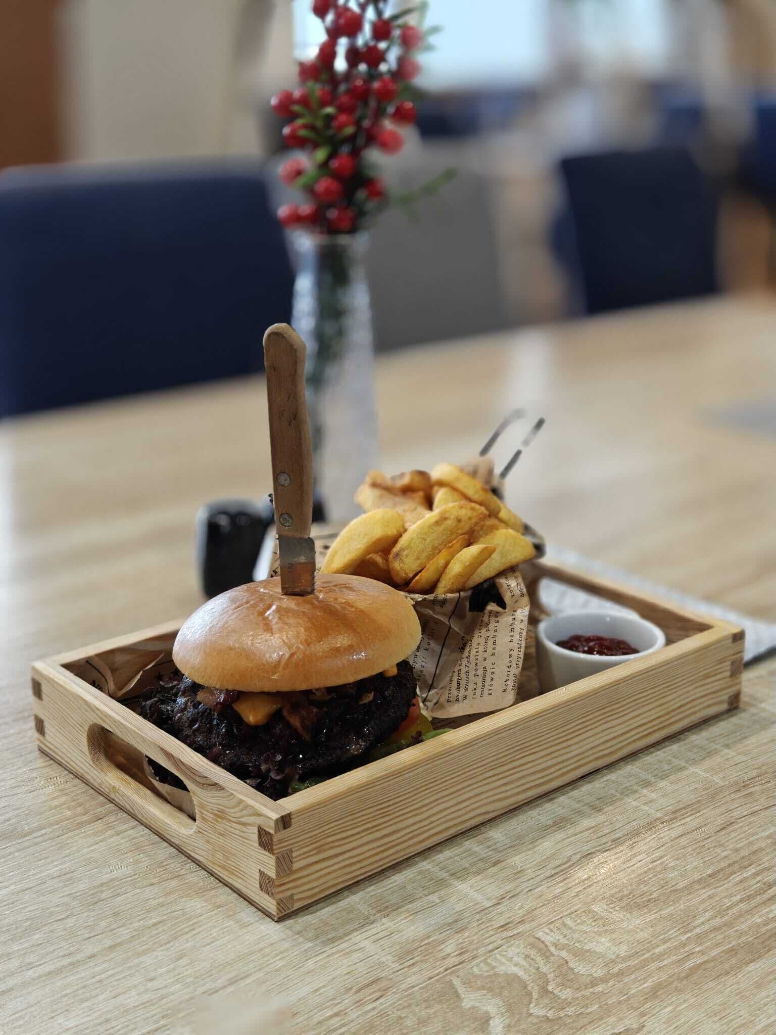 Una bandeja de madera con una hamburguesa, papas fritas y un vasito de salsa sobre una mesa.