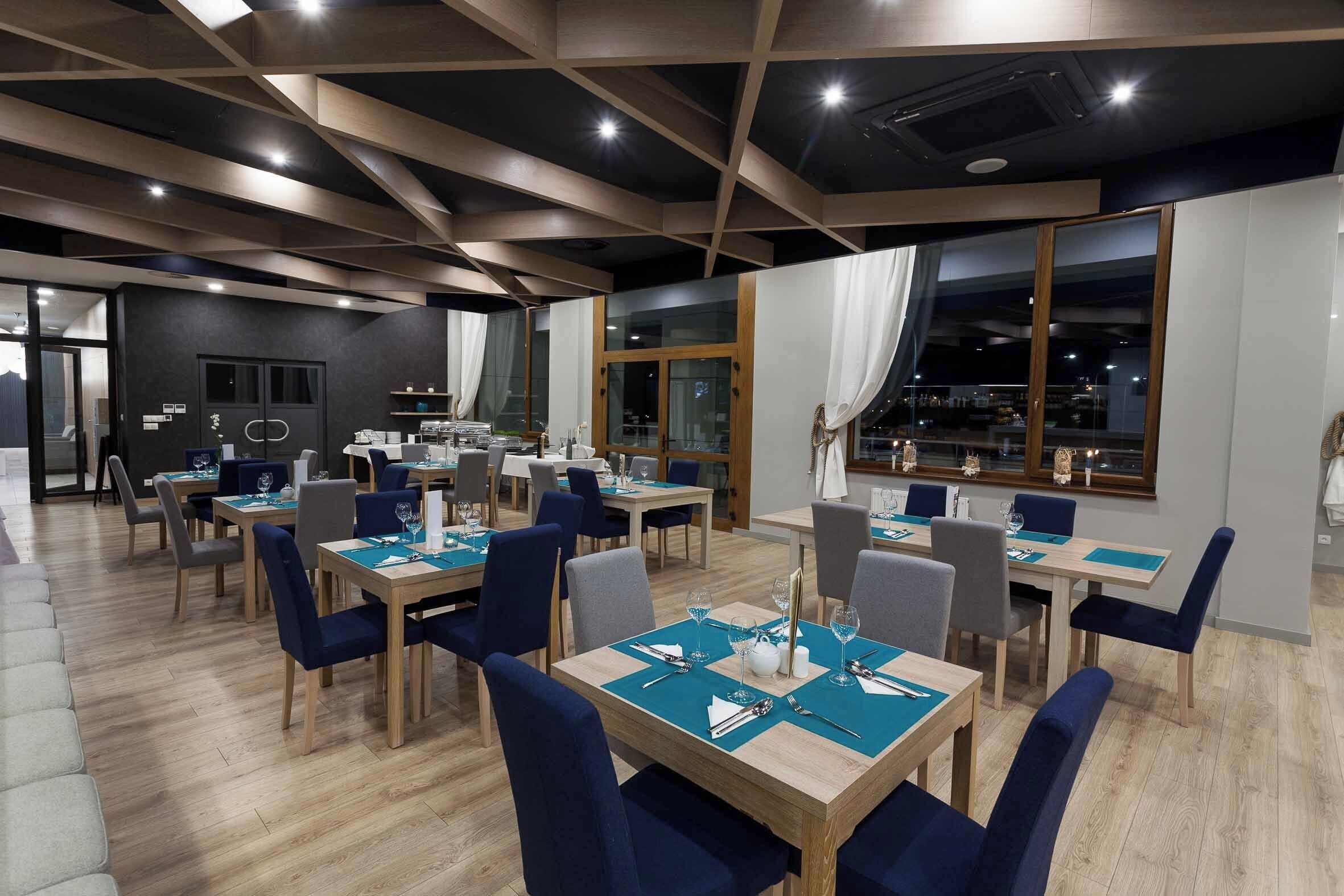 Un comedor moderno con mesas de madera y sillas azules, ventanas amplias con cortinas y un techo de madera con vigas a la vista.