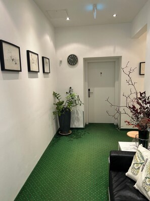 A hallway with a green patterned floor, a black sofa with a decorative pillow, a potted plant, and three framed artworks on the wall.