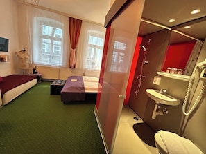 A hotel room with a bed, a small desk, a bathroom with a sink and shower, and large windows with curtains. - Pension Stadthalle (Vienna)
