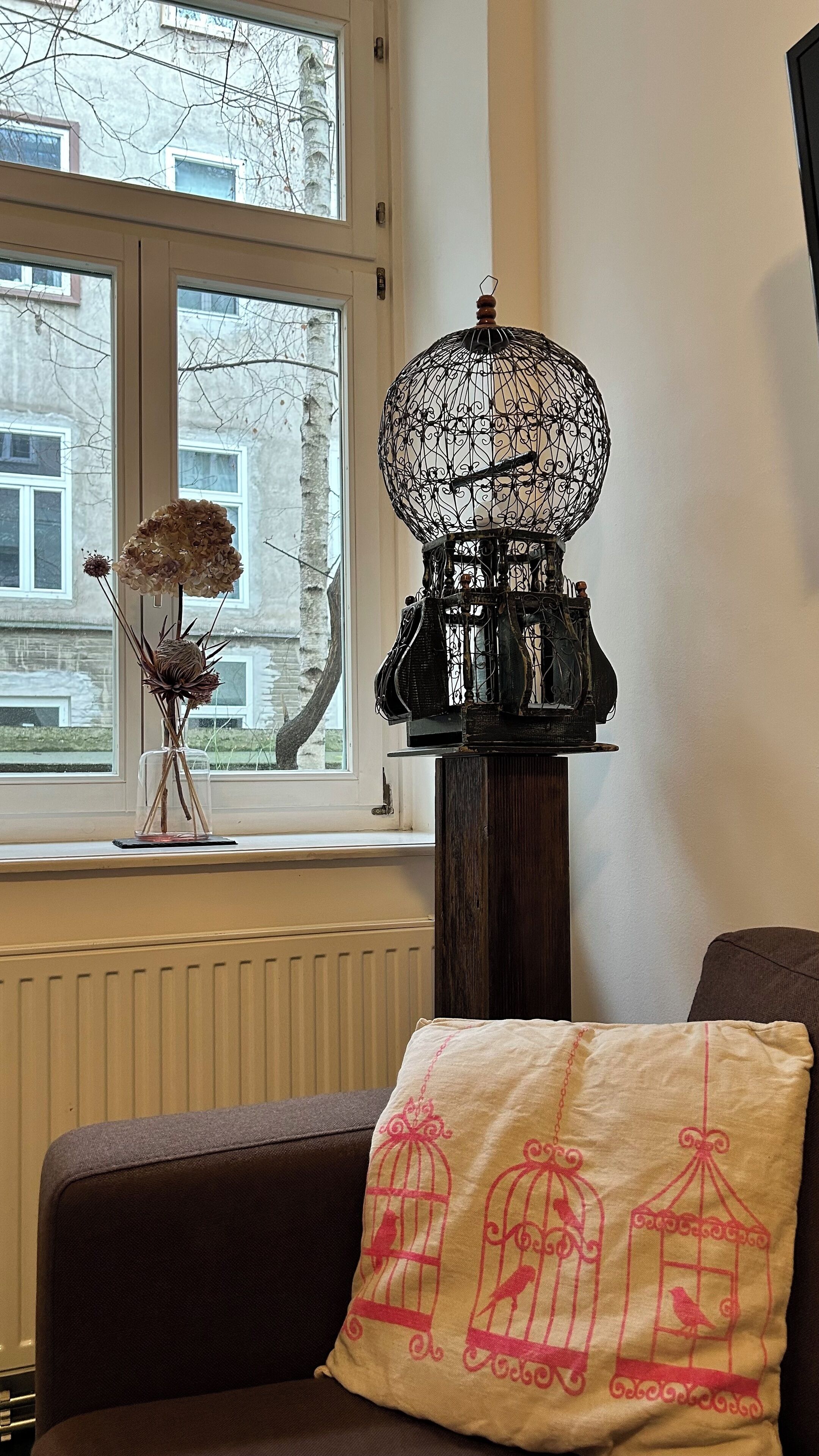 Une pièce avec une fenêtre, une lampe décorative en forme de cage à oiseaux, un vase contenant des fleurs séchées et un coussin orné de motifs de cages à oiseaux.