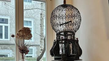 Une pièce avec une fenêtre, une lampe décorative en forme de cage à oiseaux, un vase contenant des fleurs séchées et un coussin orné de motifs de cages à oiseaux.