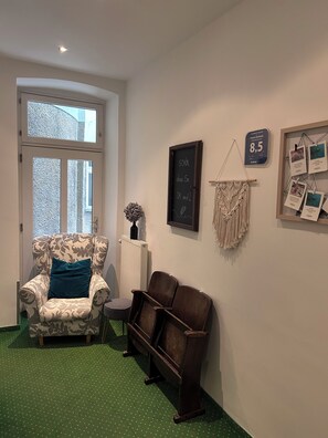 A room with a patterned armchair, a wooden bench, a small table, a radiator, a wall-mounted macrame, and a chalkboard with text.